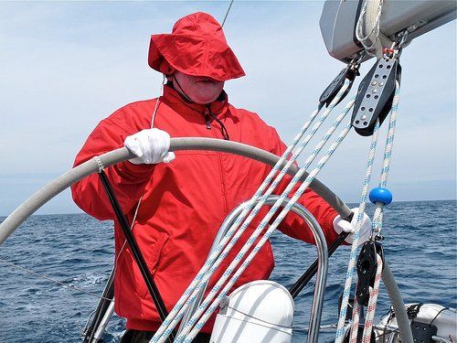 A close up image of a V.I. sailor at the helm of an underway yacht. He is wearing wet weather gear, and the wind has blown the brim of his sou'wester style hat over his face.