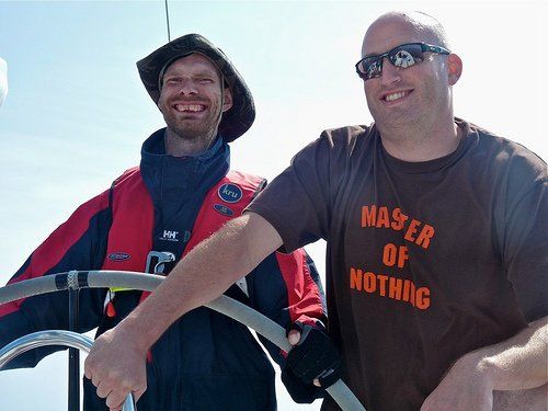 A close-up, low-angle picture of a male VI sailor and sighted male crew at the wheel of a yacht. Both are smiling. The sighted crew wears a t-shirt with the slogan Master of Nothing. It's a sunny day.