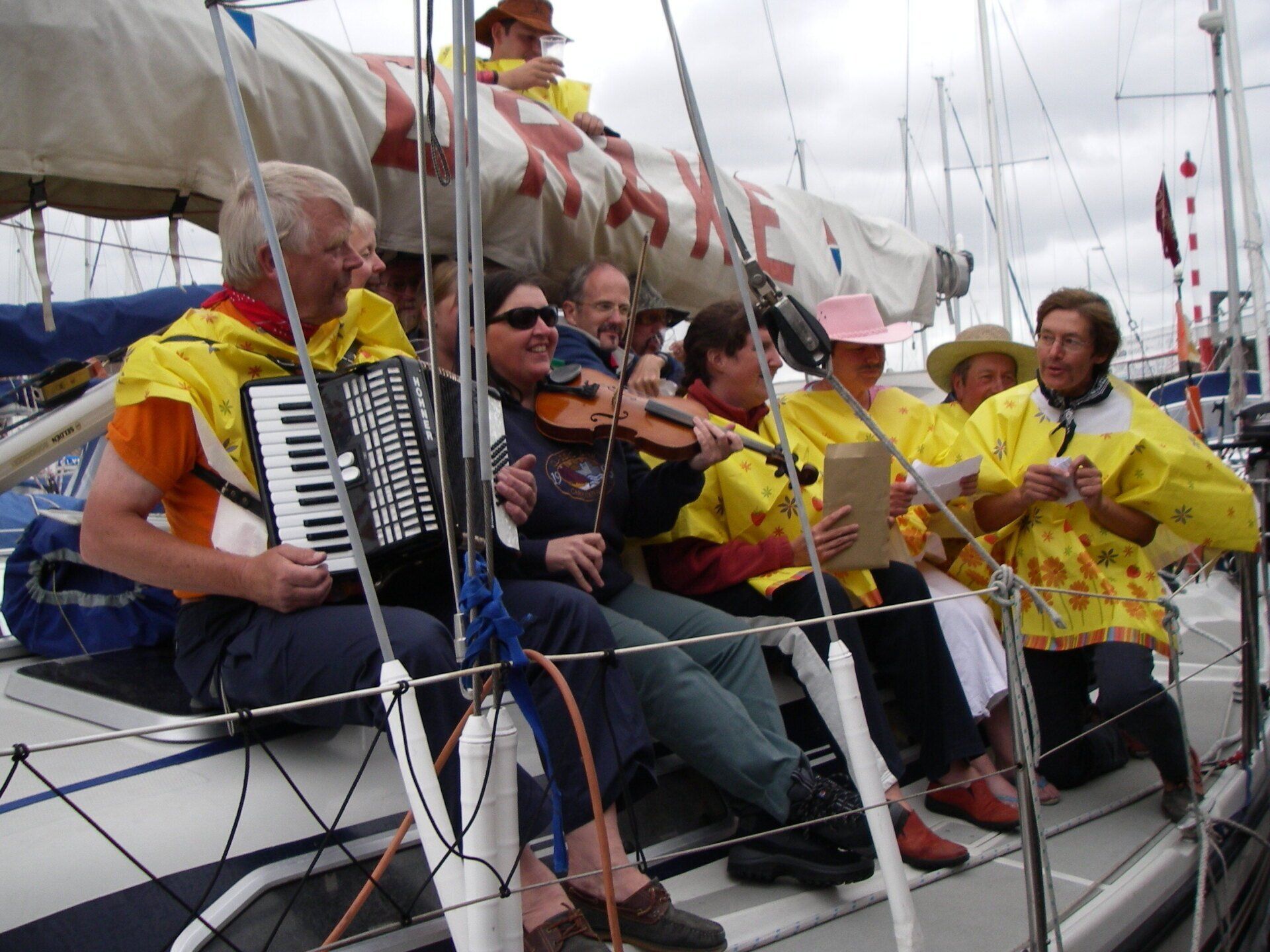 A picture taken from the pontoon in a marina. On the deck of a moored yacht a group of crew and musicians are sitting. One has an accordion, another has a fiddle and others have song sheets. They are dressed in colourful rain ponchos and some are weraing pink cowboy hats.
