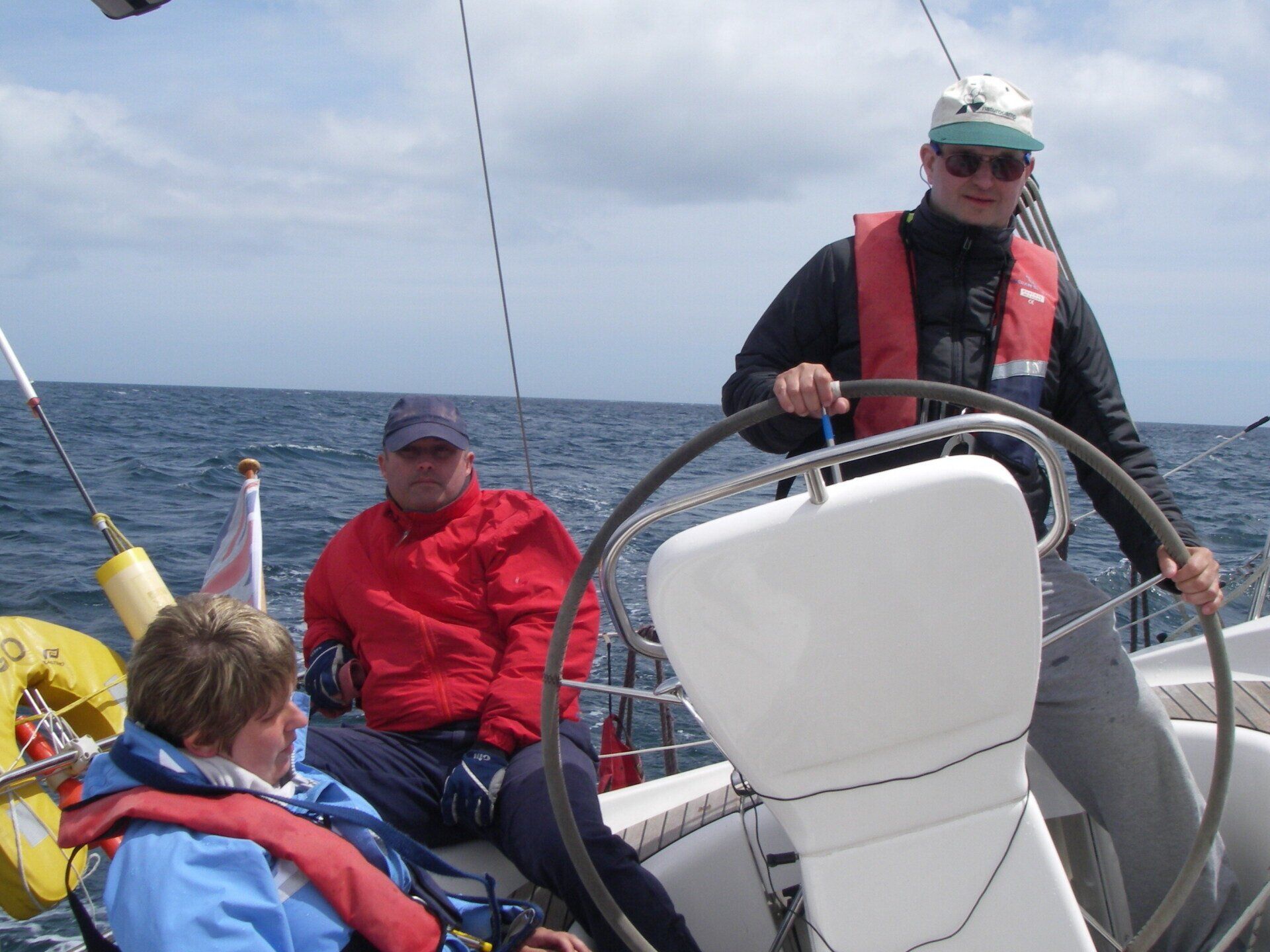 A picture of a V.I. sailor helming an underway yacht on a relatively clear day. A couple of other crew members are sitting nearby.