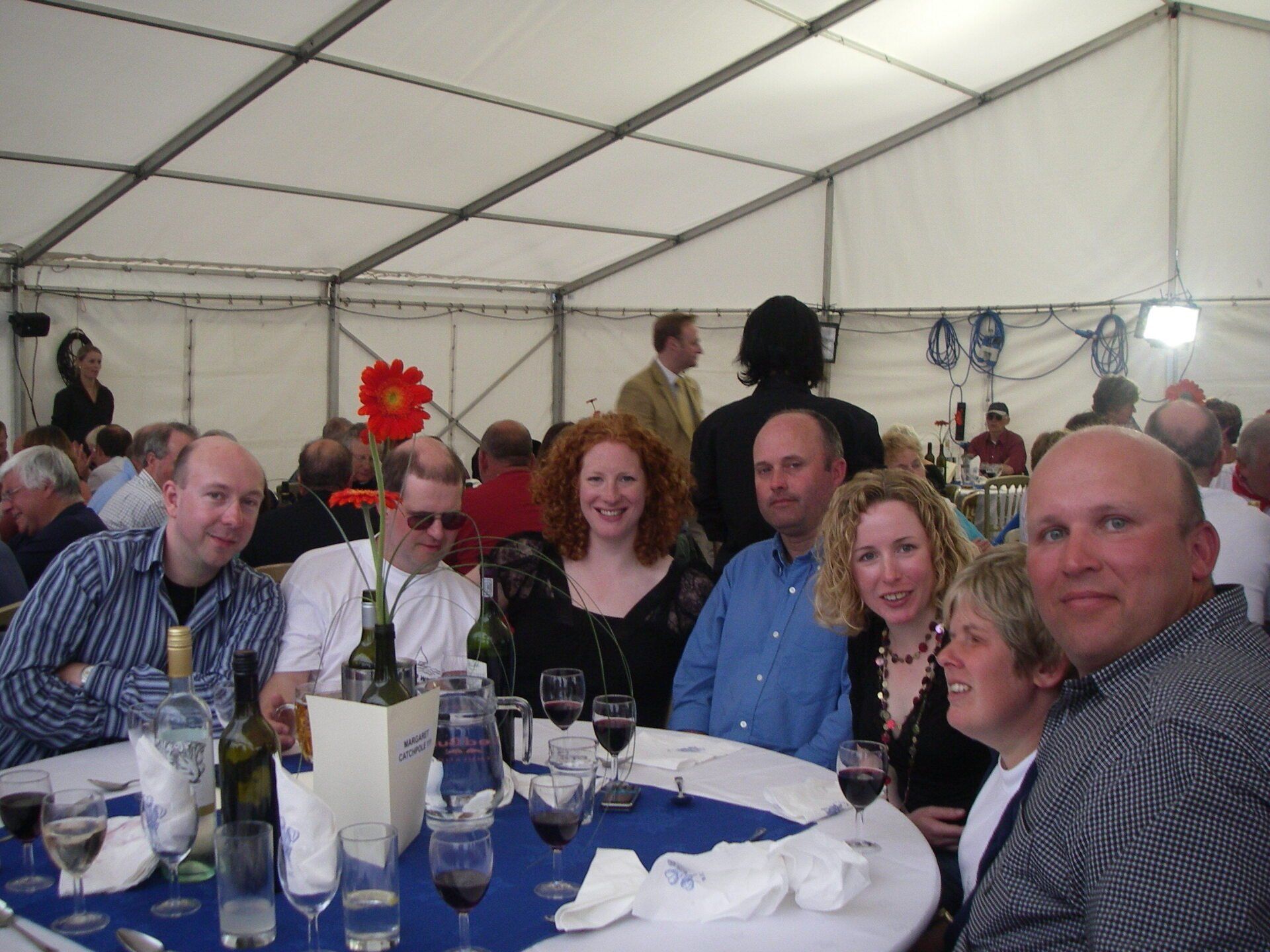 A close up picture of a smiling group sitting around a dining table in a marquee. Other tables and groups are in the background. Glasses of wine and used napkins on the table indicate that this is the end of dinner.