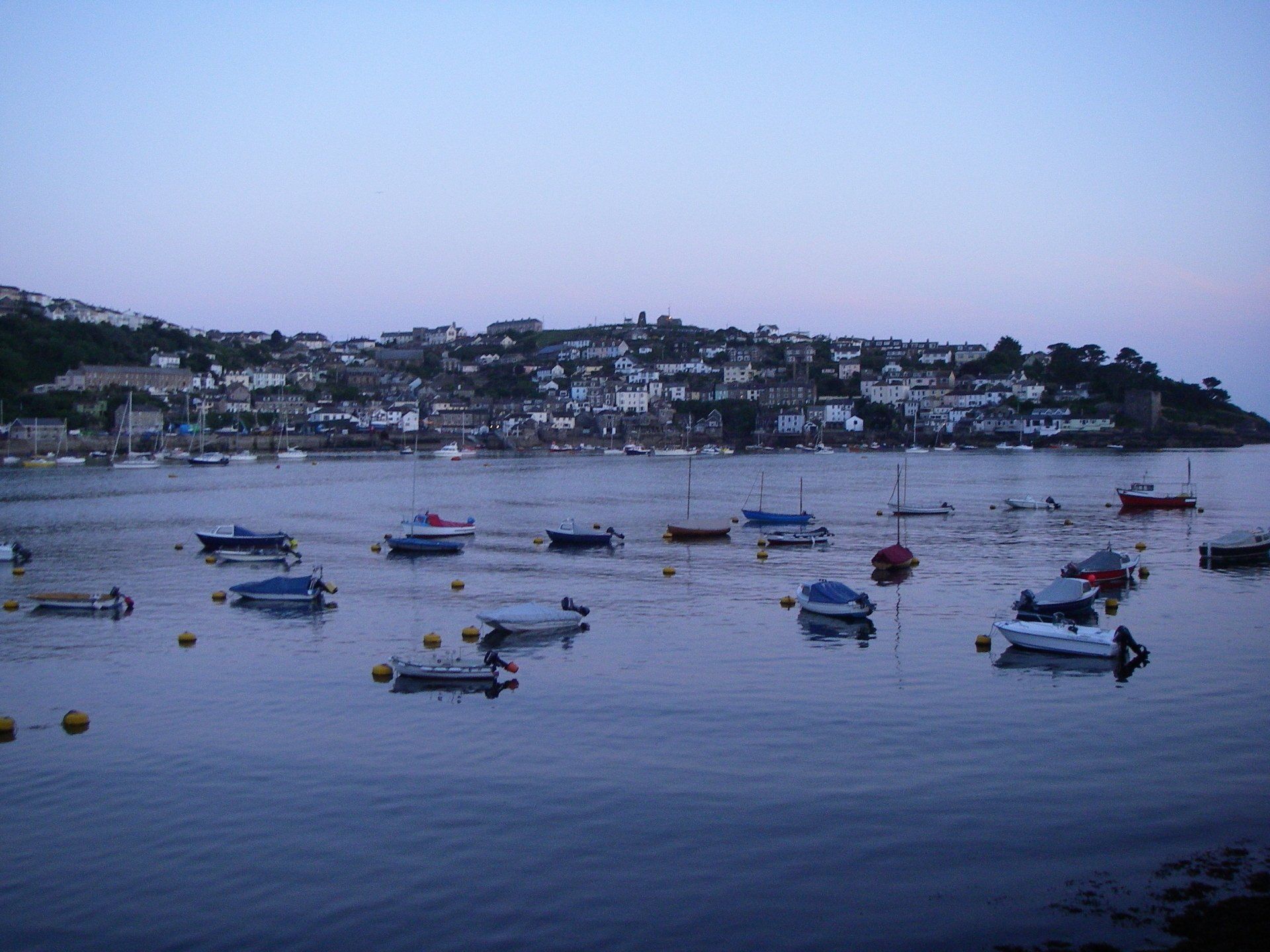 A landscape and seascape picture taken in the evening. The foreground is a small bay in which several boats and yachts are moored. In the background is a coastal town - possibly Falmouth? It is dusk and the sea is very calm.