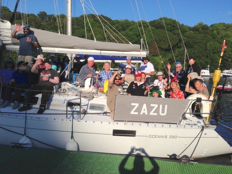 A picture taken from a pontoon of a large group of V.I. and sighted sailors sitting on the deck of the yacht 'Zazu', which is moored. The crew are raising glasses. It's a sunny evening, with the shadow of the photographer visible in the foreground.