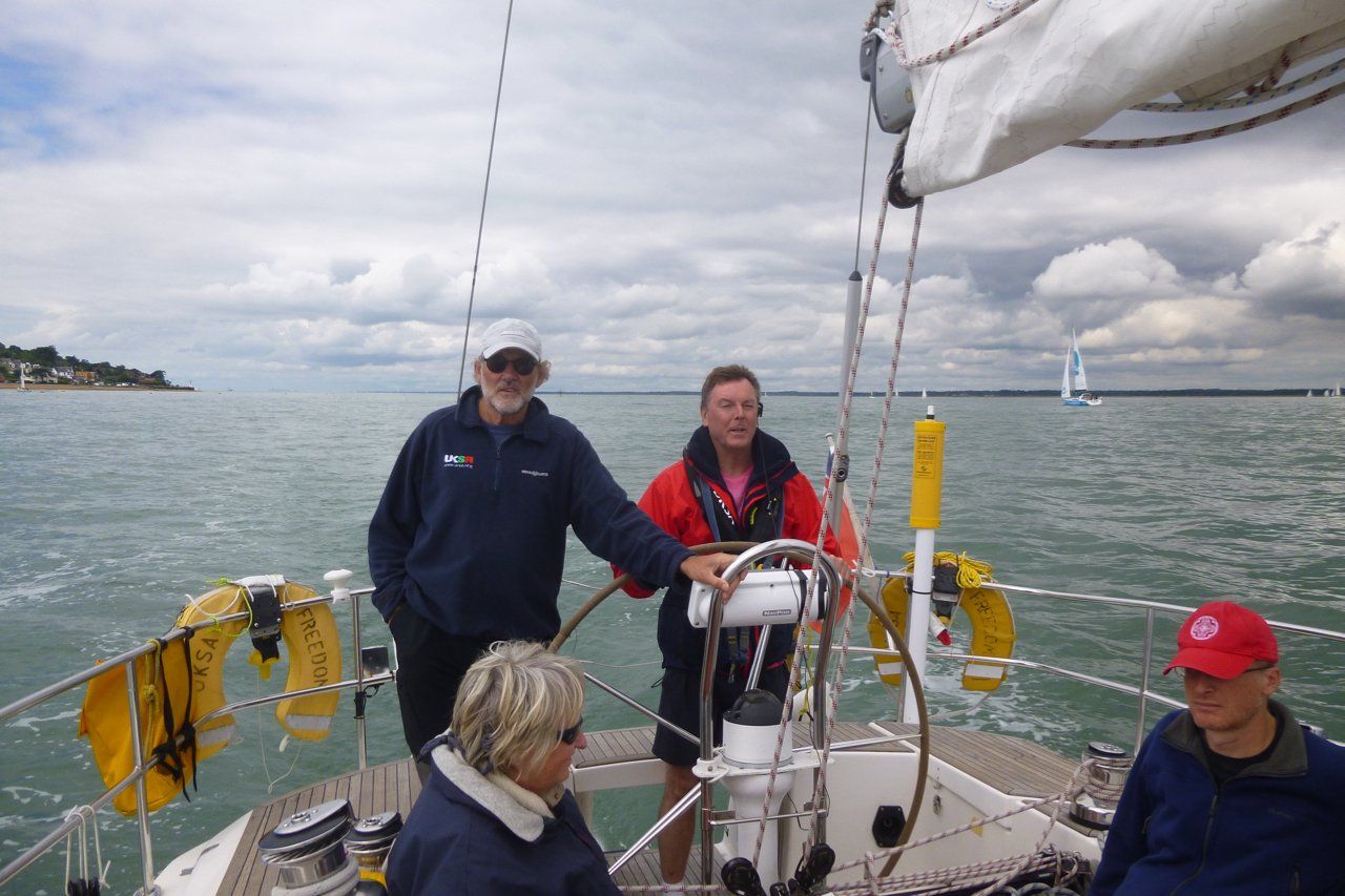 A picture taken from the deck of a yacht under sail, looking back at the wheel. A V.I. sailor is at the helm, with sighted and V.I. crew sitting and standing nearby. Conditions are overcast but calm.
