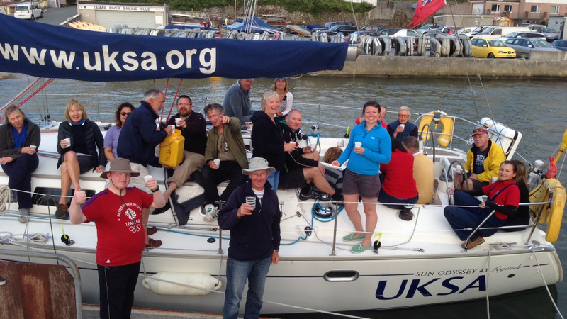 A picture taken at the end of a day's sailing of around twenty sighted and V.I. crew on the deck and adjacent pontoon. The group are smiling and raising glasses to the camera.