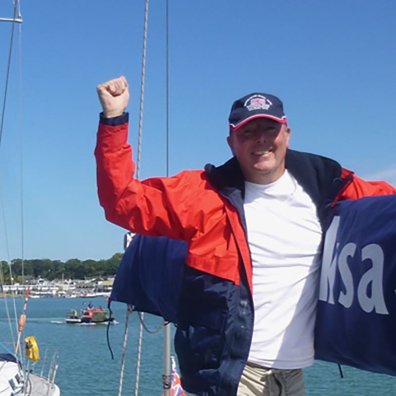 A close up of a V.I. sailor making his way along the deck of a yacht. One arm is over the boom, the other raised in the air in a cheering motion. He is smiling broadly at the camera.