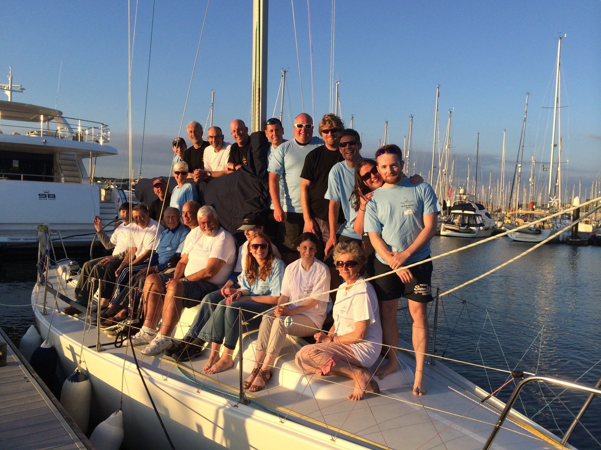 A group picture of nineteen V.I. and sighted crew, sitting and standing on the deck of a yacht moored at a marina pontoon. It is evening and the sun is shining.