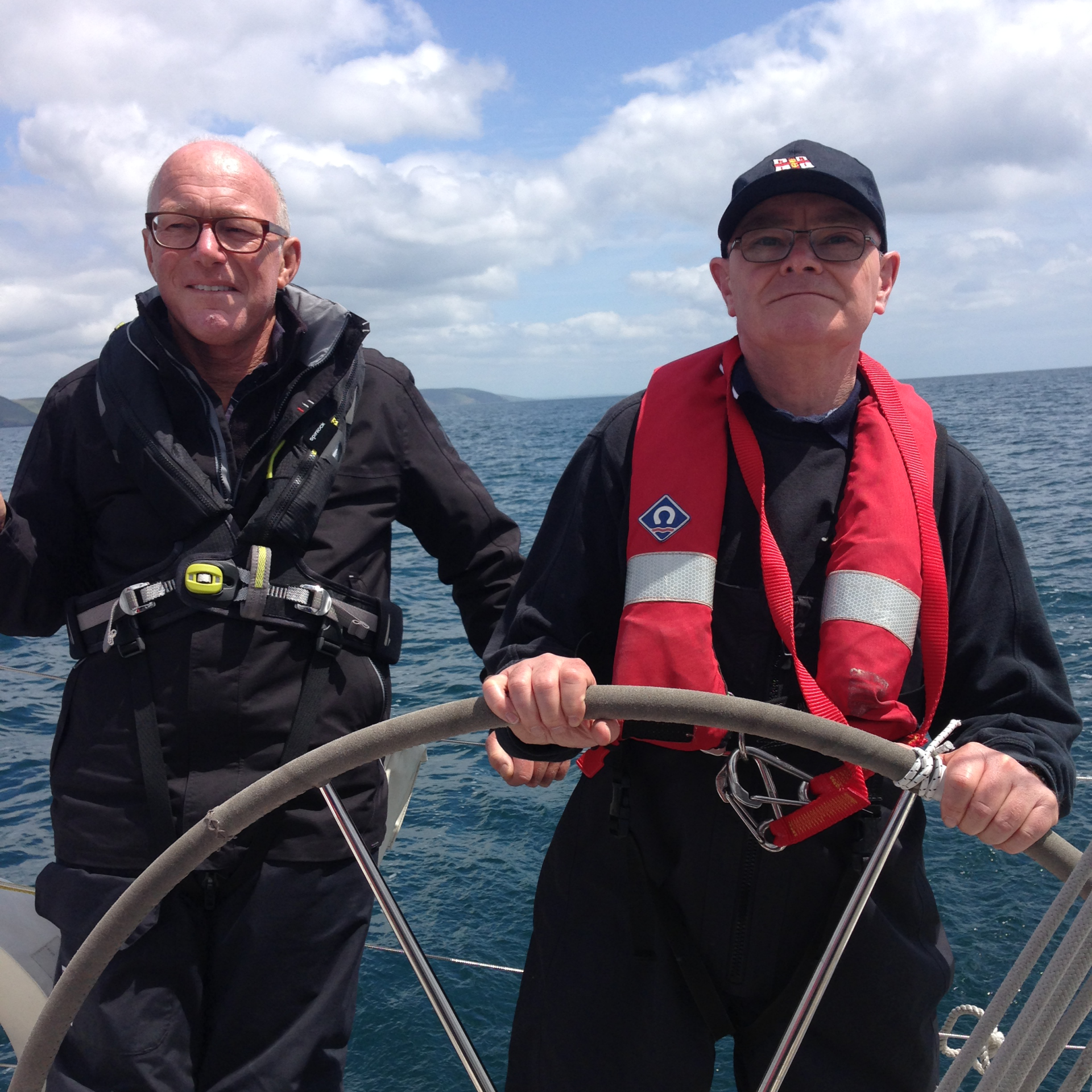 A picture taken looking back at the wheel of a yacht under sail. It is a clear, bright and calm day. A V.I. sialor is at the helm, with a sighted crew member assisting.