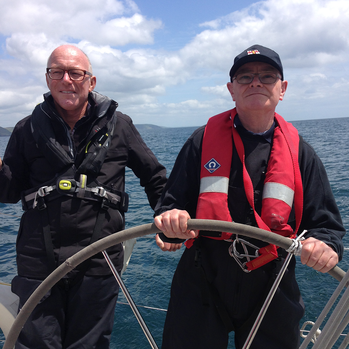 A picture of a sighted male skipper standing next to a male VI sailor who has the wheel. The sea is calm.