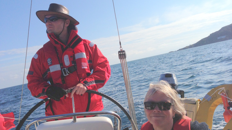 A picture taken from the deck of an underway yacht, looking back to the wheel, being helmed by a visually impaired male sailor. A female sighted crew member sits next to him. The sea and horizon in the background are on an angle, showing the vessel is under sail.