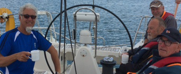 A picture of four male crew members, VI and sighted, sitting on the benches around a yacht's wheel, smiling and enjoying a cup of tea.