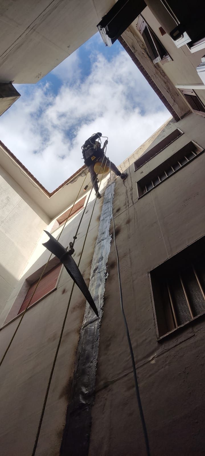 Persona che si cala in corda doppia da un edificio, vista dal basso, con cielo e nuvole visibili.