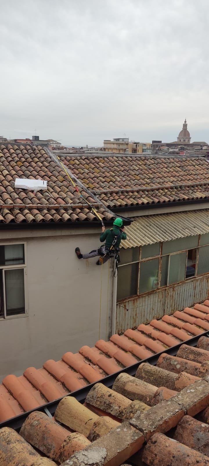 Persona in abito verde appesa al bordo di un edificio, probabilmente al lavoro. Scenario: tetto, cielo nuvoloso.