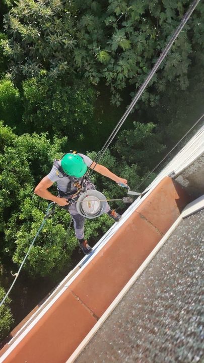 Uomo con equipaggiamento di sicurezza che si cala in corda doppia da un edificio, casco verde, attrezzatura in mano, vista dall'alto.