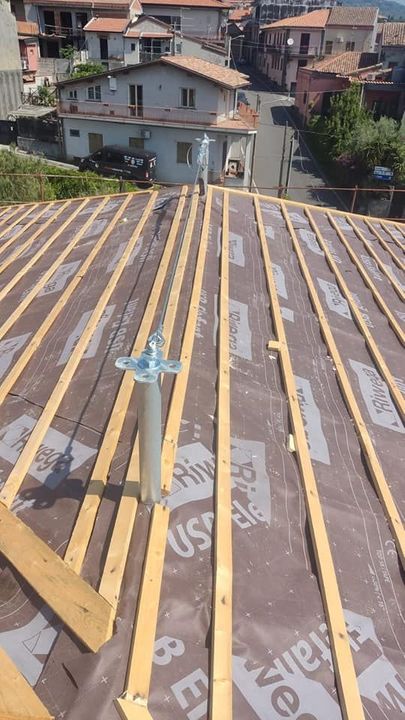 Costruzione del tetto: travi in legno e strato di impermeabilizzazione. Un tubo di sfiato si estende verso l'alto. Case e strada visibili sullo sfondo.