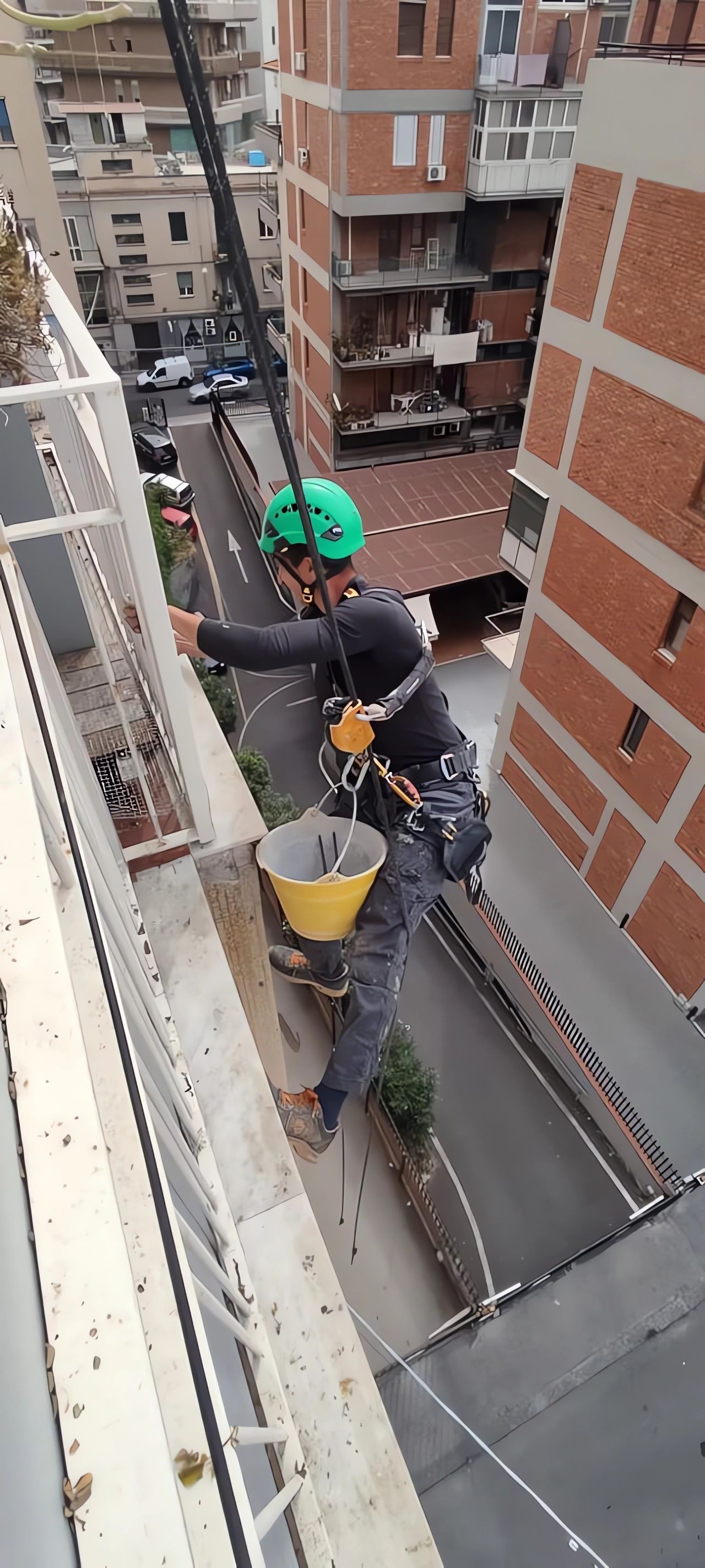 Persona che si cala in corda doppia dalla facciata di un edificio, tenendo in mano un secchio. Indossa dispositivi di sicurezza, mentre lavora su un edificio.