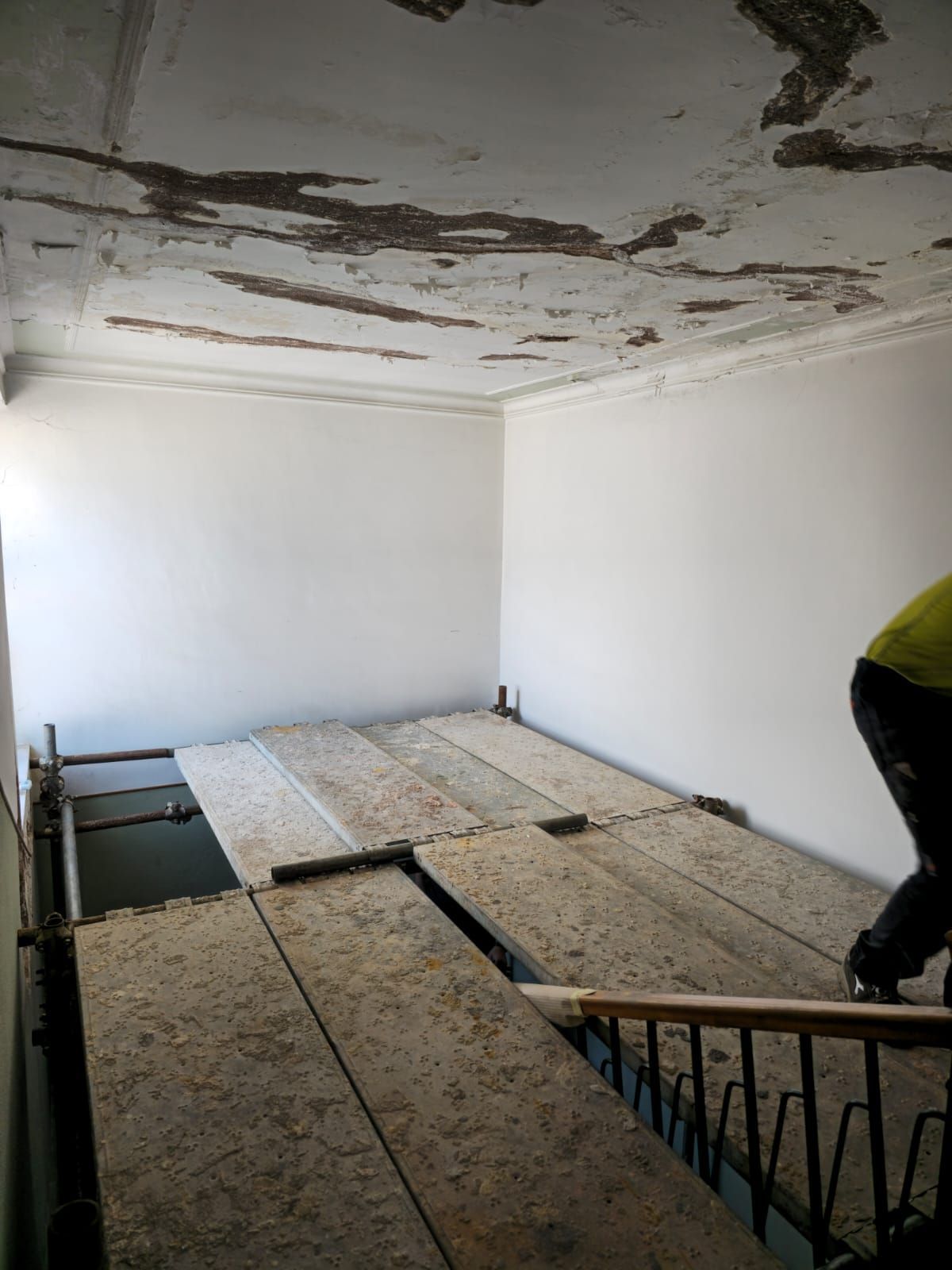 Stanza in fase di ristrutturazione con impalcature, pavimento in compensato, soffitto danneggiato, pareti bianche e una persona in un angolo.