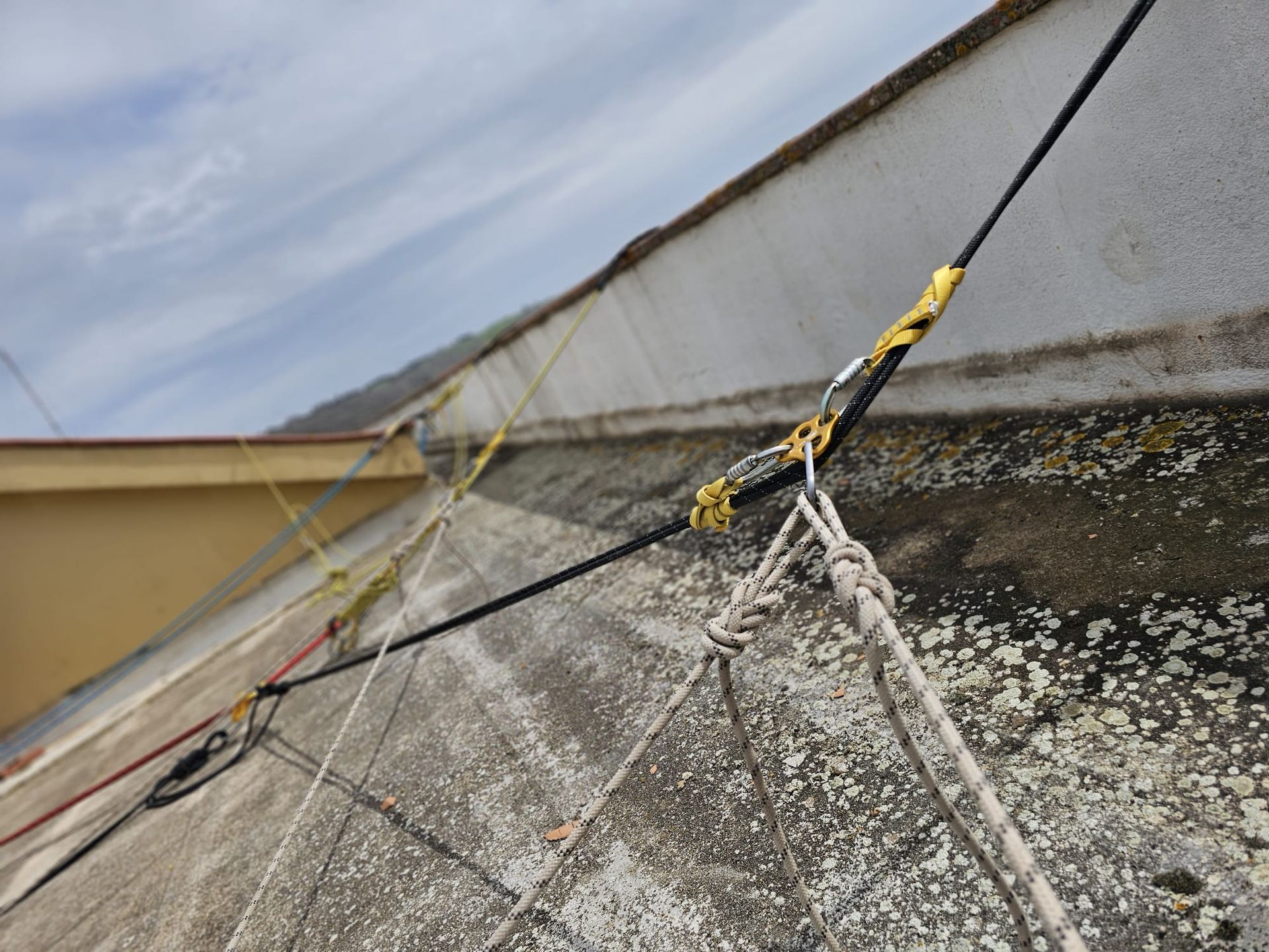 Corda nera legata a un tetto, assicurata da un moschettone e altre corde; ambiente esterno, cielo nuvoloso.