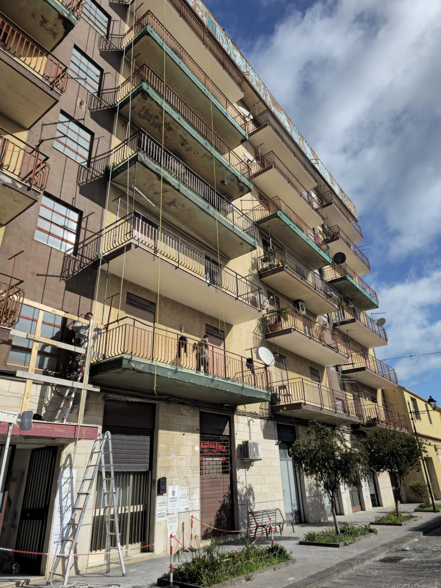 Edificio a più piani con balconi, probabilmente in fase di ristrutturazione. Esterno marrone chiaro, ringhiere in metallo, cielo azzurro.