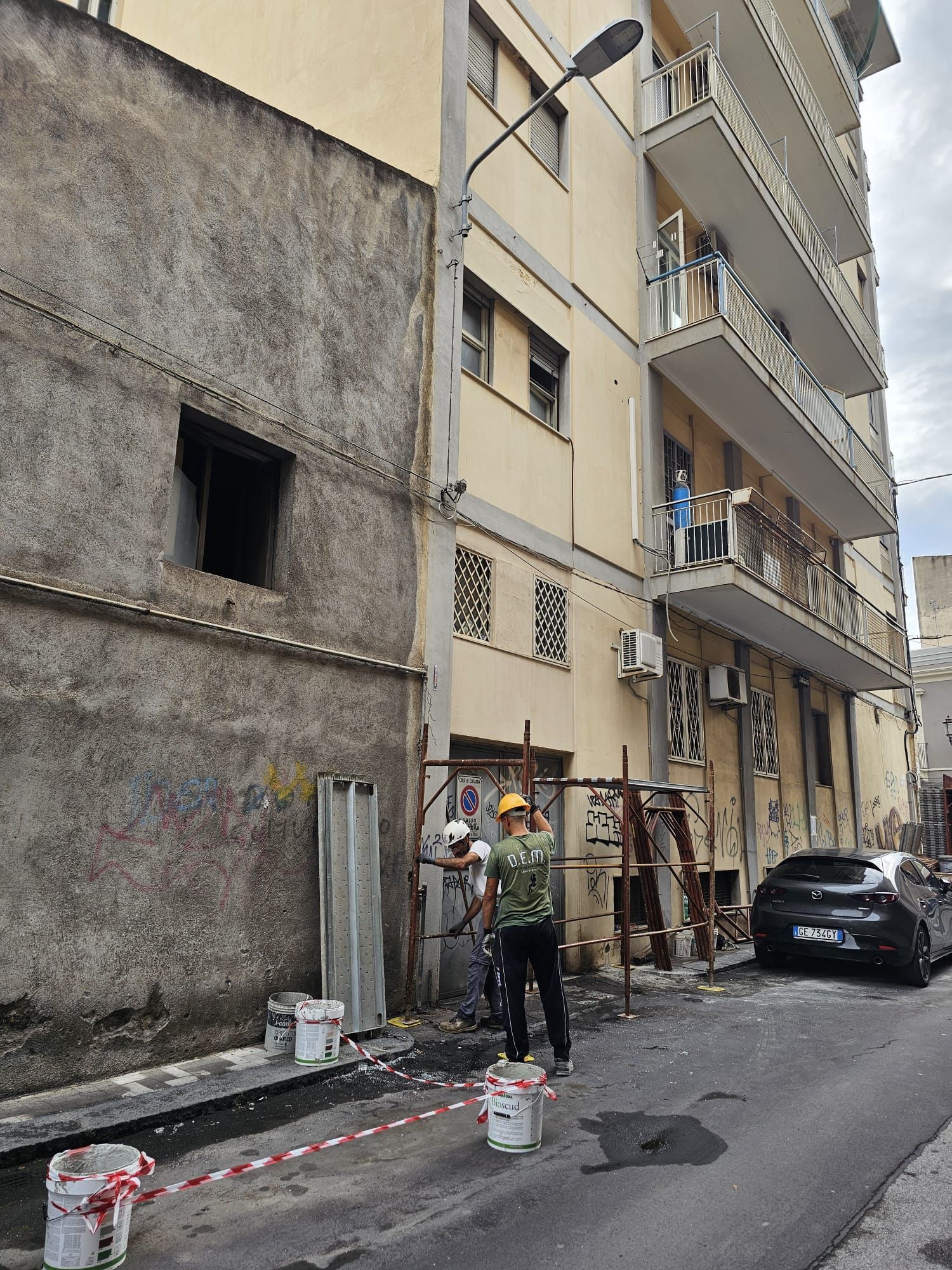 Operai che dipingono l'esterno di un edificio in una strada stretta, con un'auto parcheggiata nelle vicinanze.