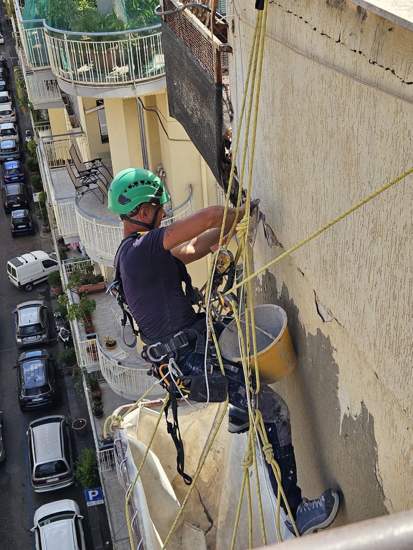 Addetto all'accesso su fune con maglietta verde e casco su un edificio, mentre installa qualcosa vicino a una finestra. Sullo sfondo della città.