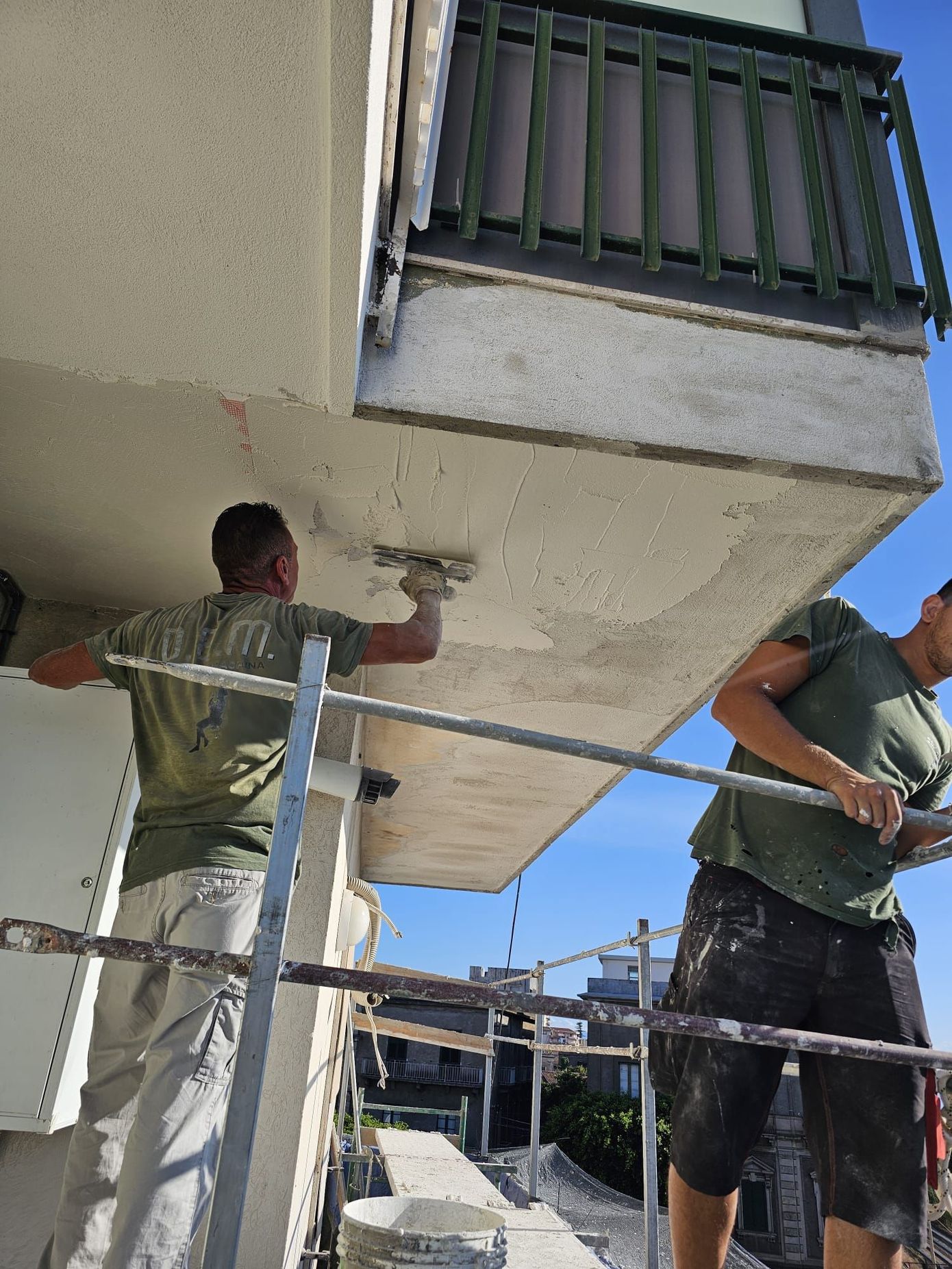 Due uomini stanno intonacando il soffitto di un balcone; uno è su un'impalcatura.
