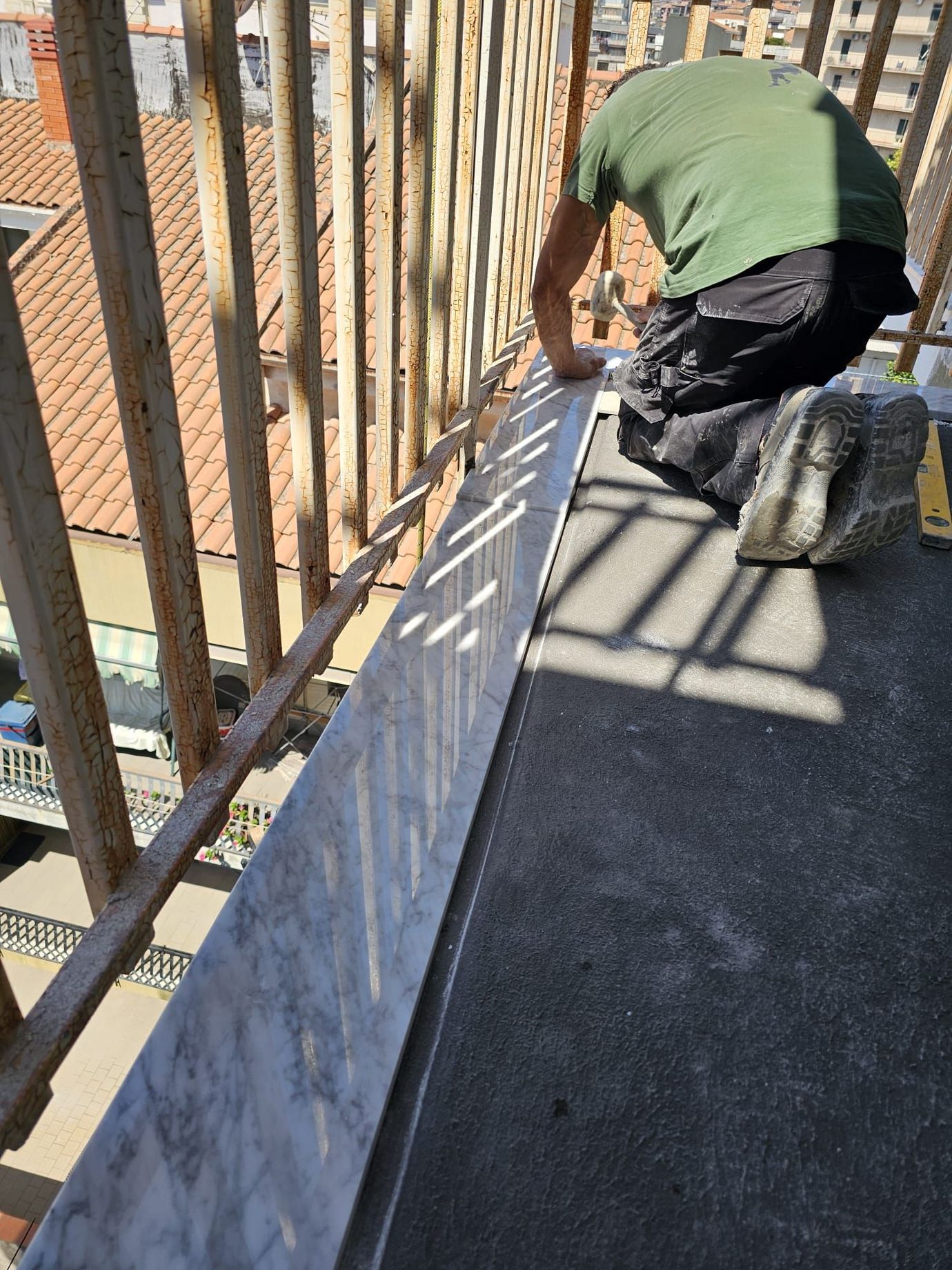 Una persona inginocchiata su un balcone installa delle finiture in marmo, indossando una maglietta verde e pantaloncini neri, vicino a una ringhiera.