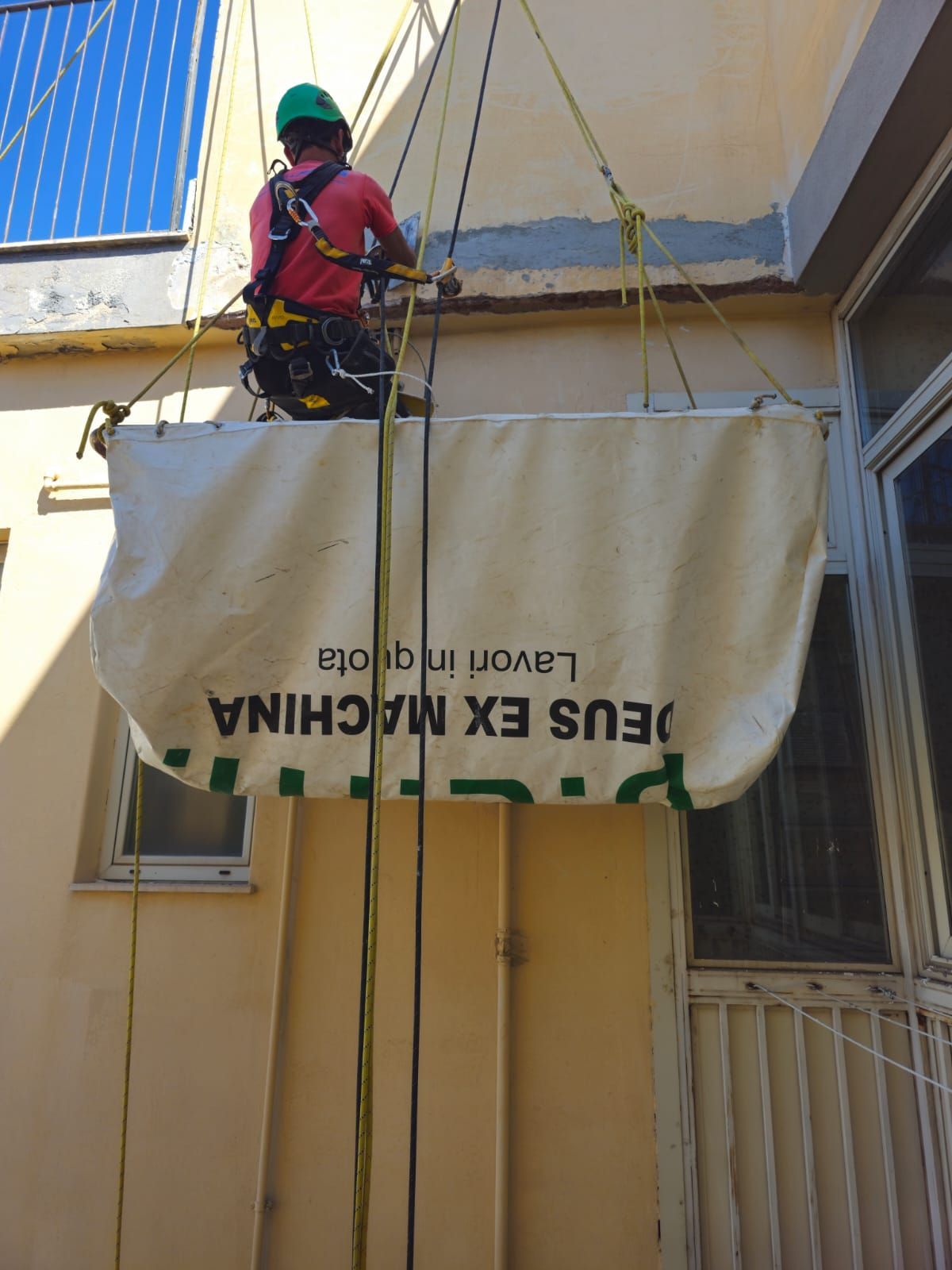 Operaio imbracato sospeso su un edificio, che maneggia un grosso sacco con la scritta