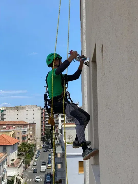 Addetto all'accesso su fune con maglietta verde e casco su un edificio, mentre installa qualcosa vicino a una finestra. Sullo sfondo della città.