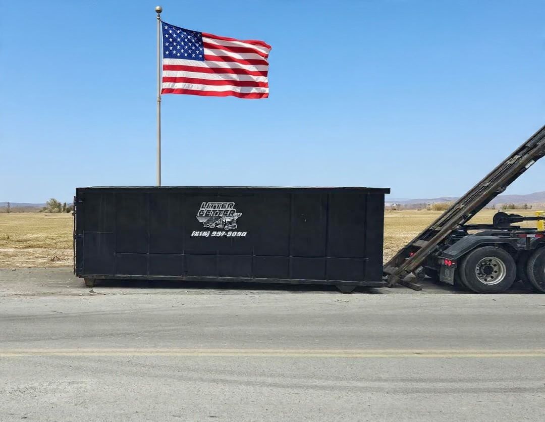 Black dumpster on a truck bed with a waving American flag against a clear blue sky.