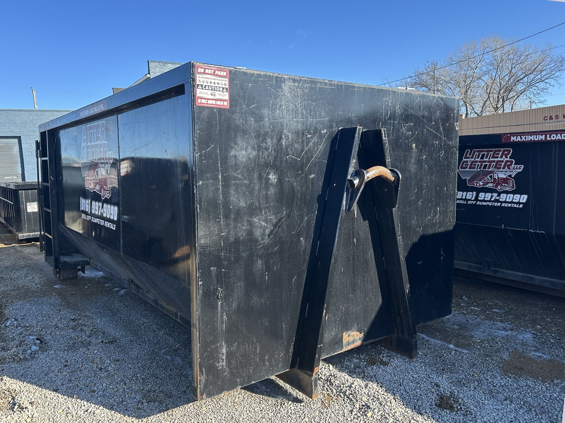 Black dumpster on gravel with a lifting mechanism.