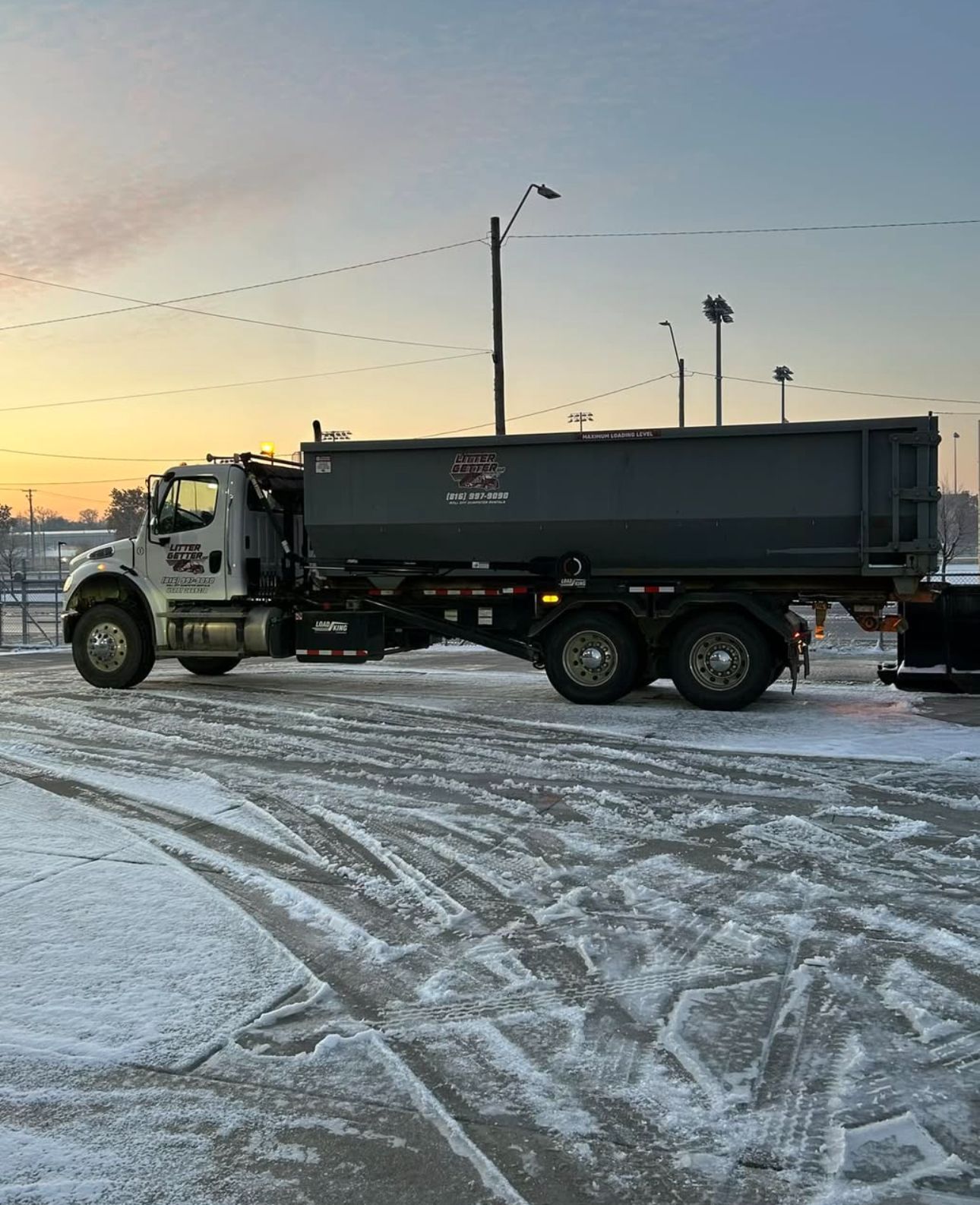 Commercial Roll-off dumpster truck parked on a snowy lot. Grey container, white cab, tire tracks.