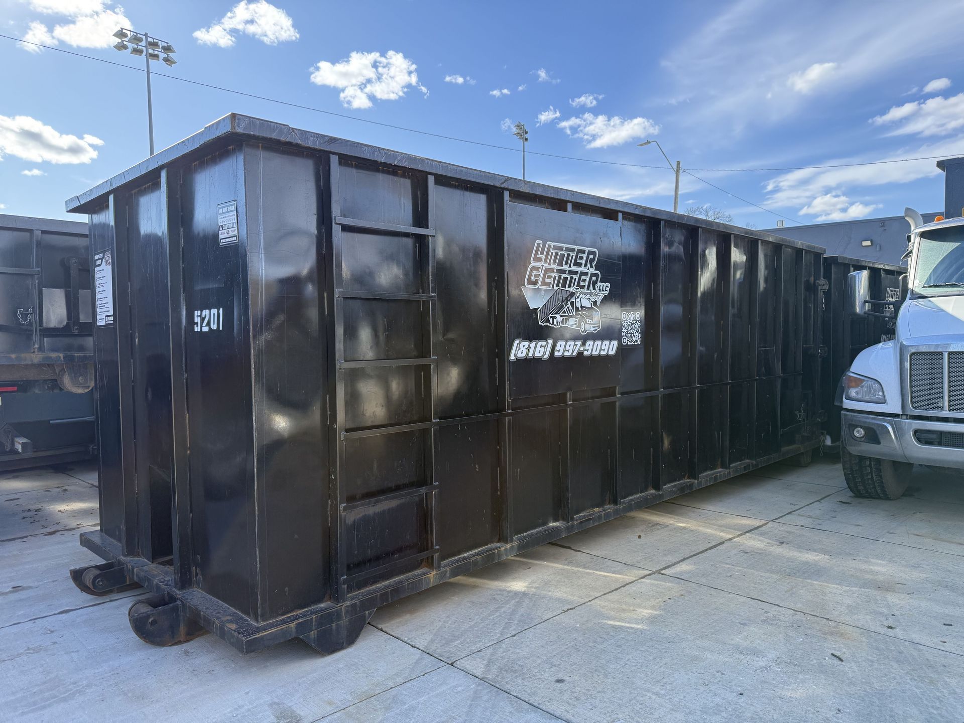Black metal dumpster trailer parked on a concrete lot under a blue sky, with a truck beside it.