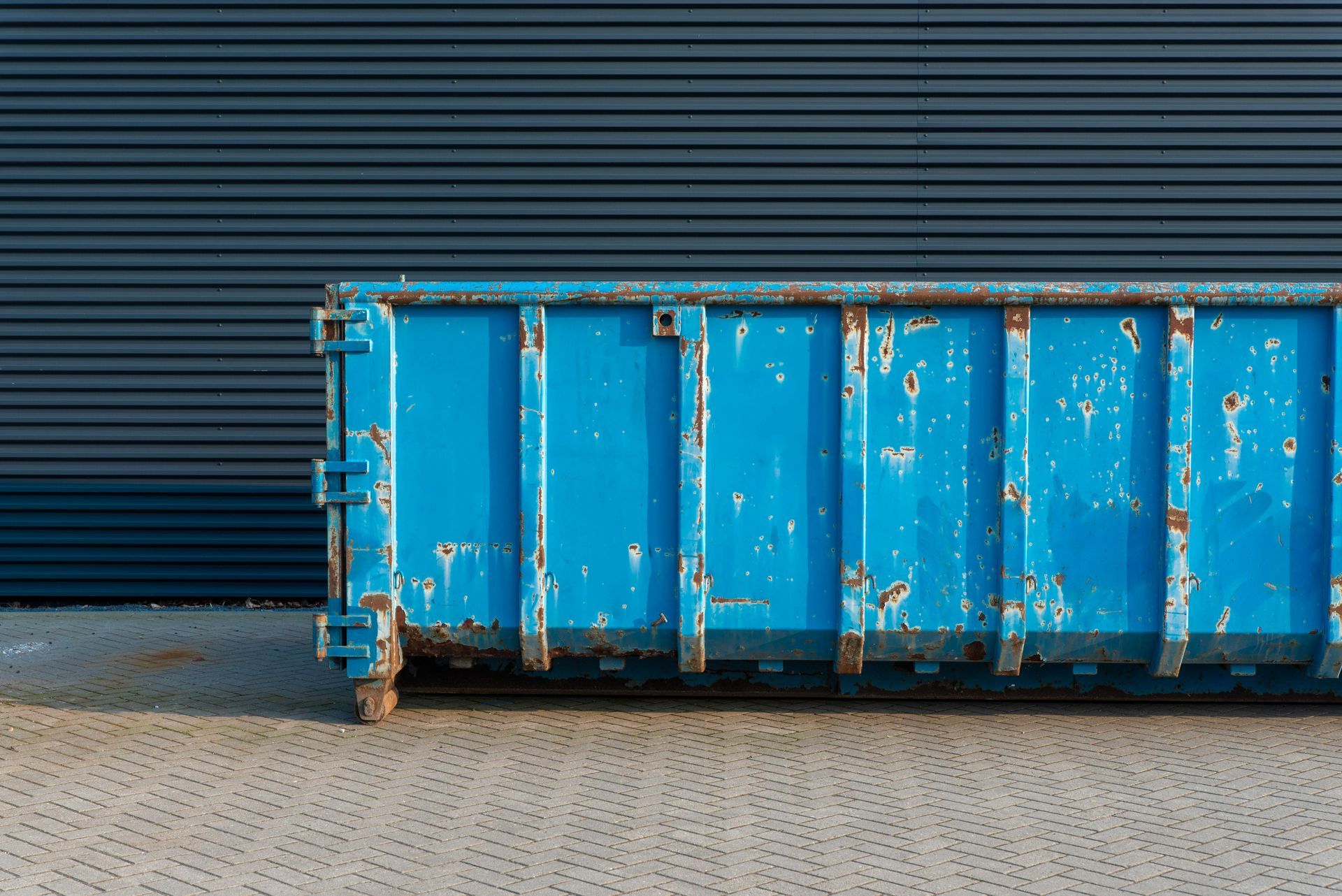 A weathered blue dumpster sits on a textured pavement in front of a dark corrugated metal wall.