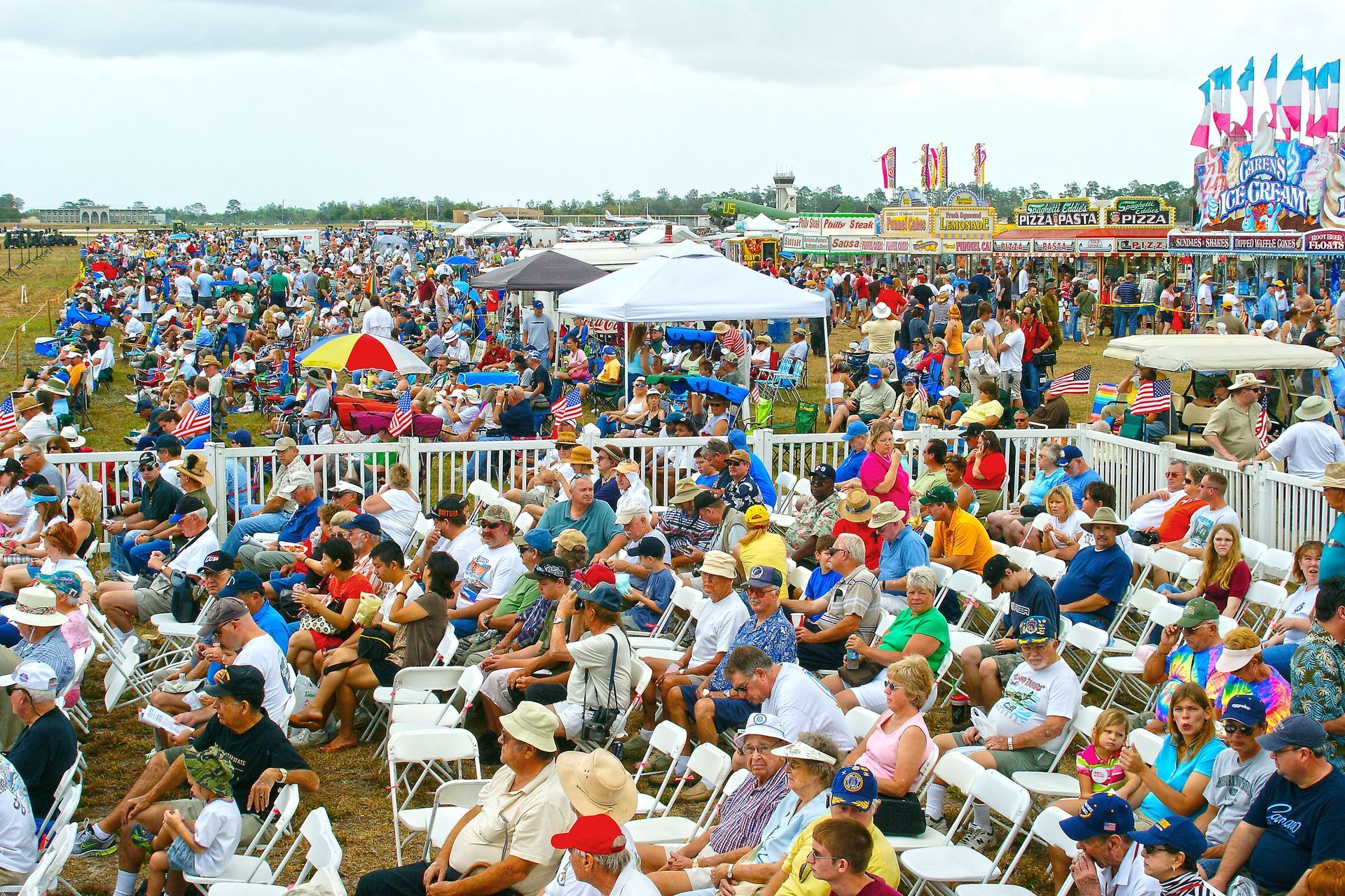 Large crowd seated outdoors at a colorful event, with tents and flags in the background.