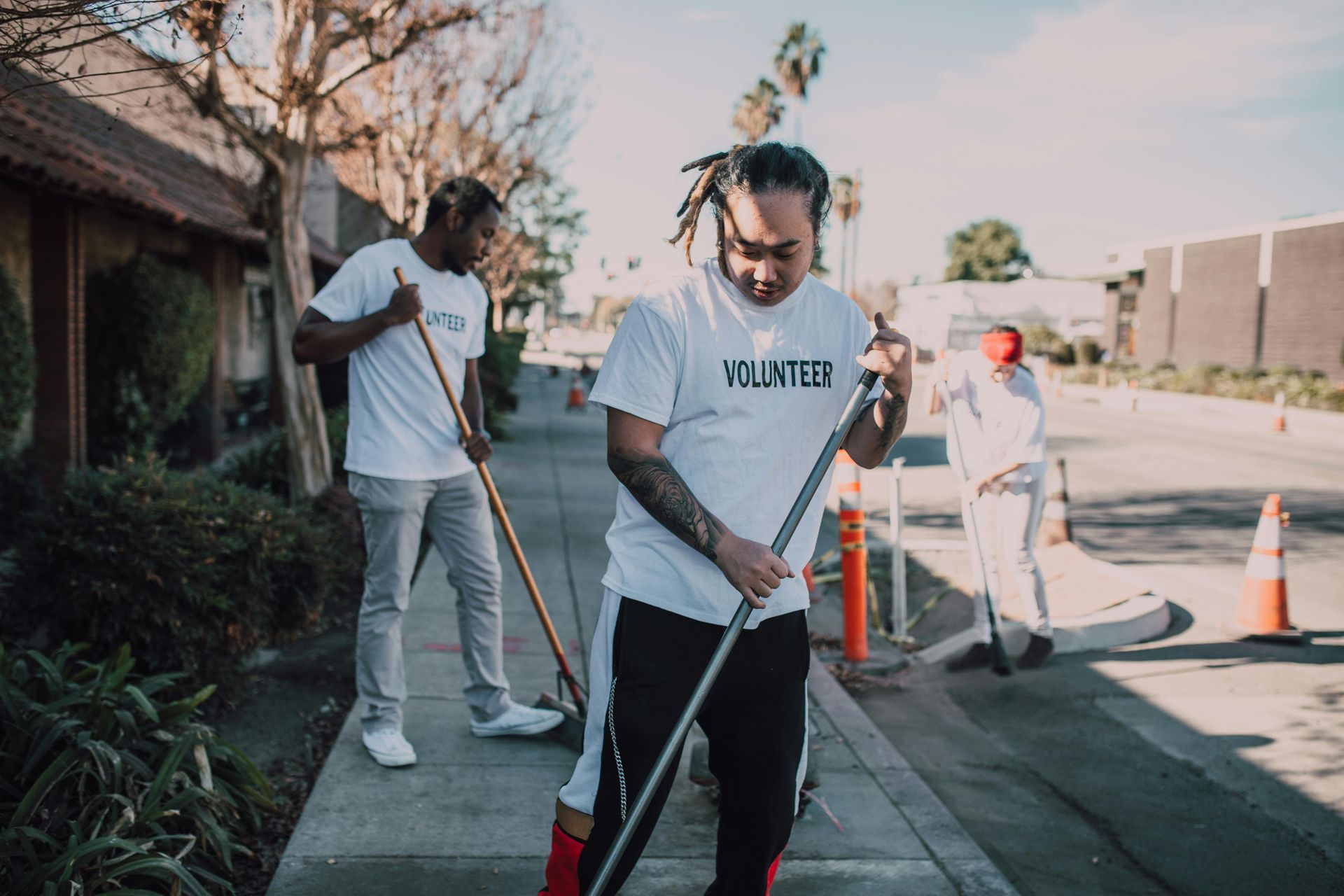 Three volunteers wearing white shirts are sweeping a sidewalk along a road on a sunny day.