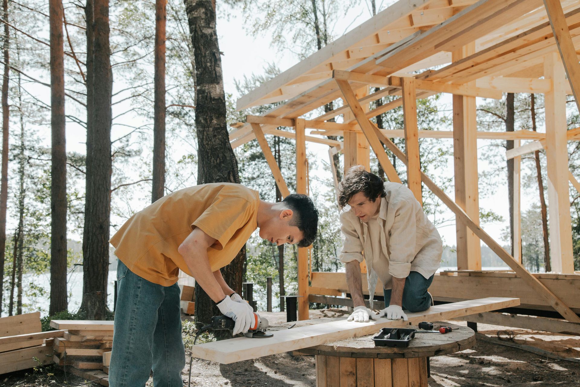 Two people building a wooden structure outdoors. One saws wood while the other kneels, working.