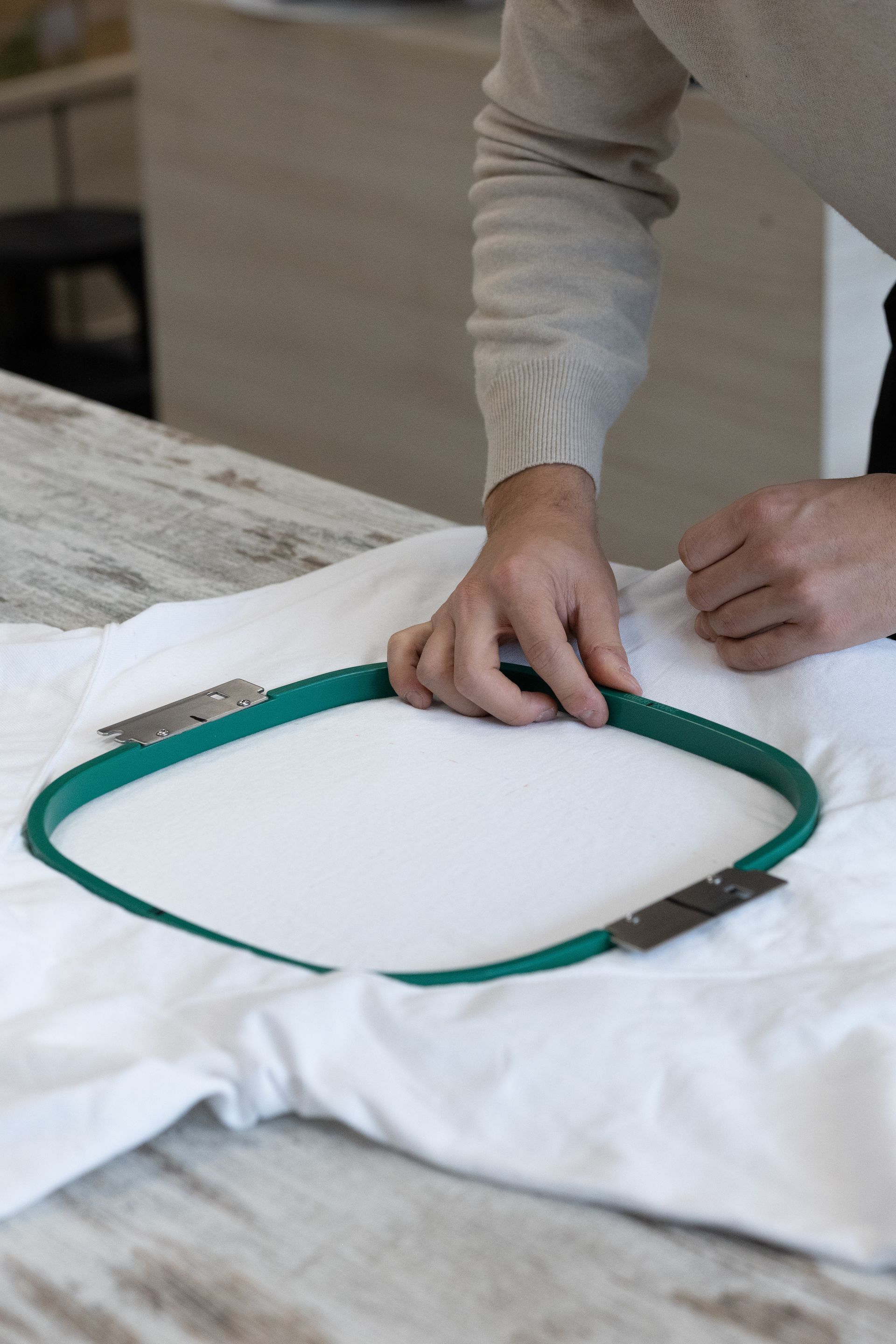 Persona che posiziona un telaio da ricamo quadrato verde su una maglietta bianca posta su un tavolo.