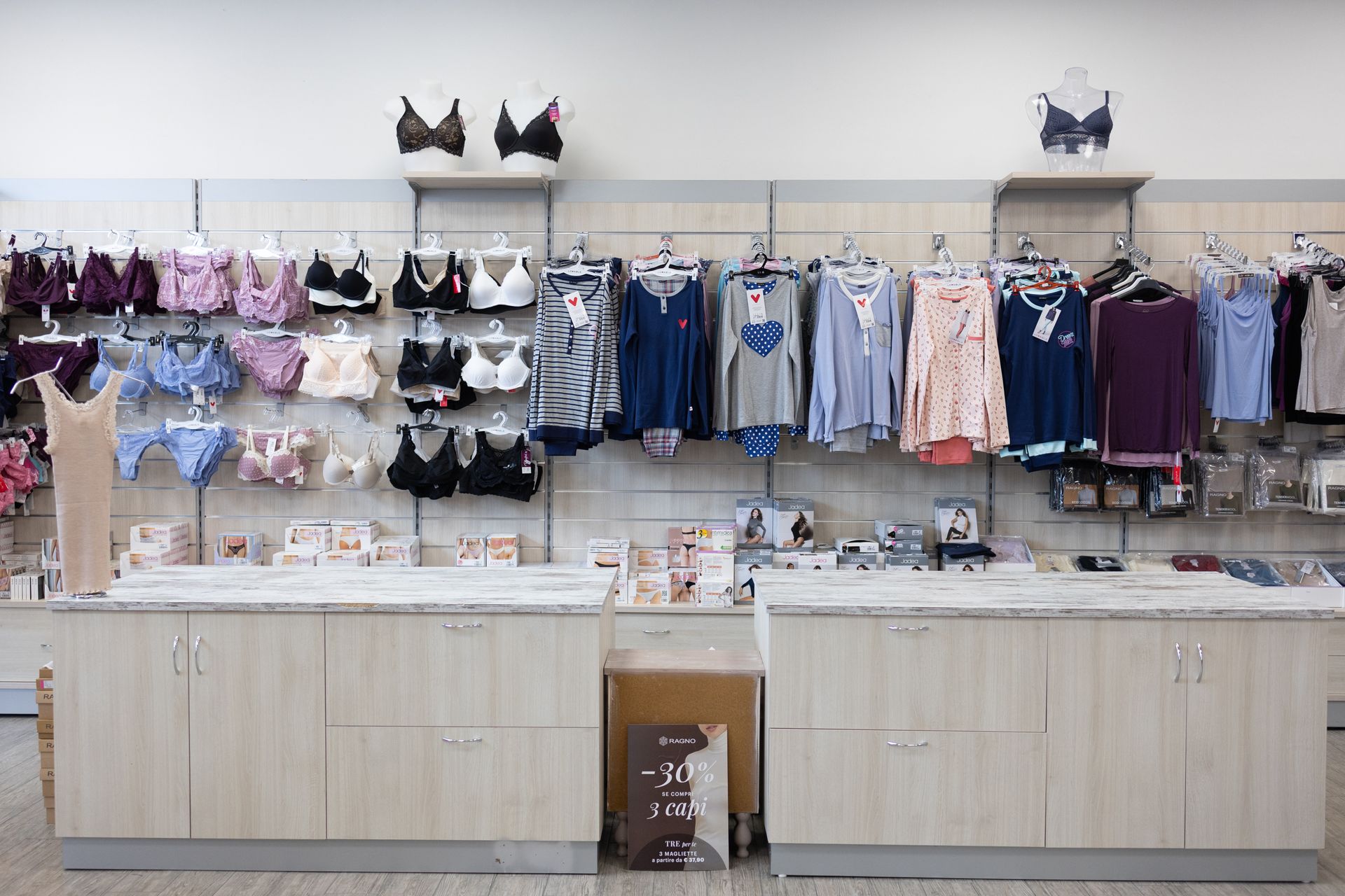 Interno di un negozio di abbigliamento con reggiseni e top esposti su scaffali, ripiani beige sottostanti.