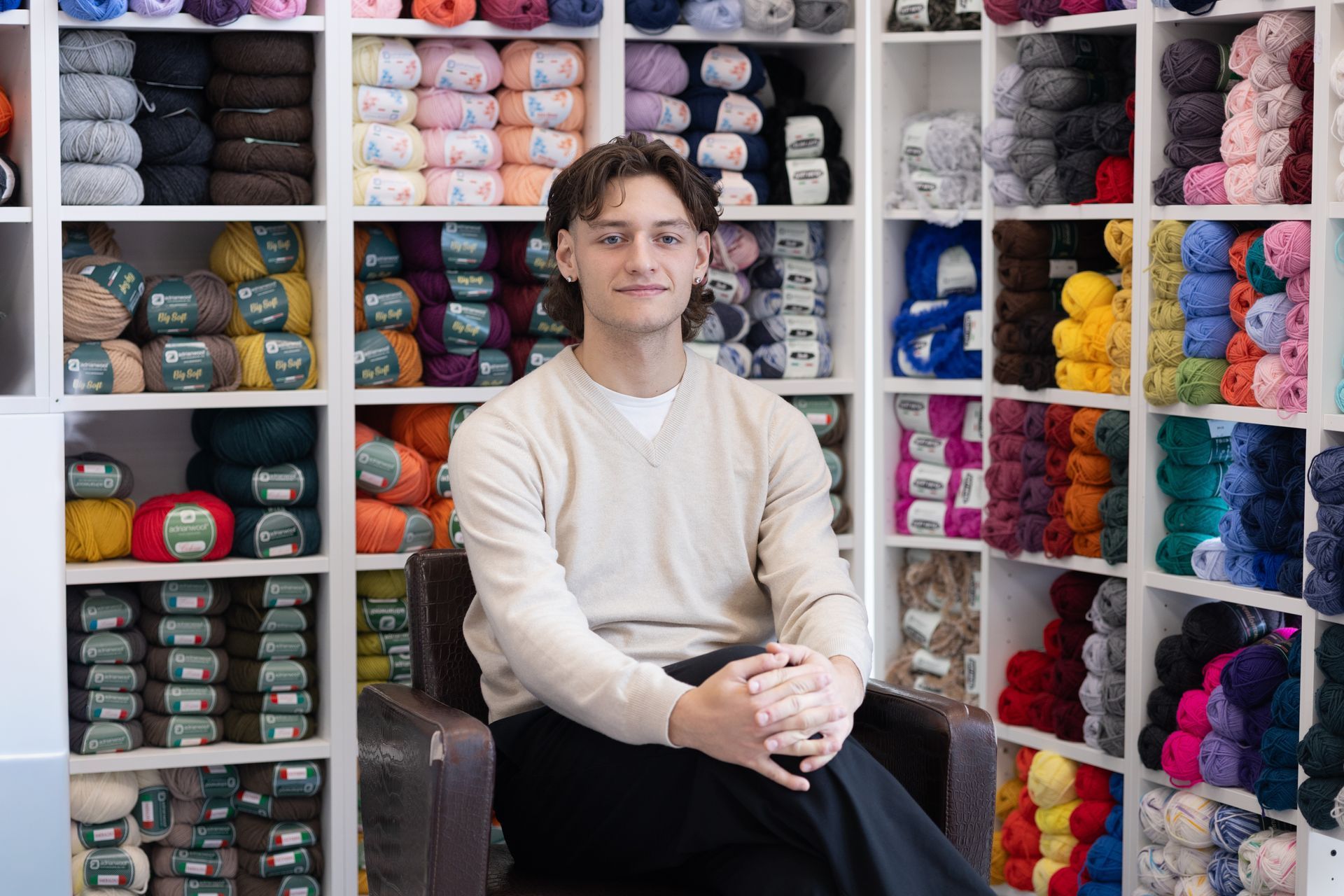 Uomo seduto in un negozio di filati, circondato da scaffali pieni di filati colorati.