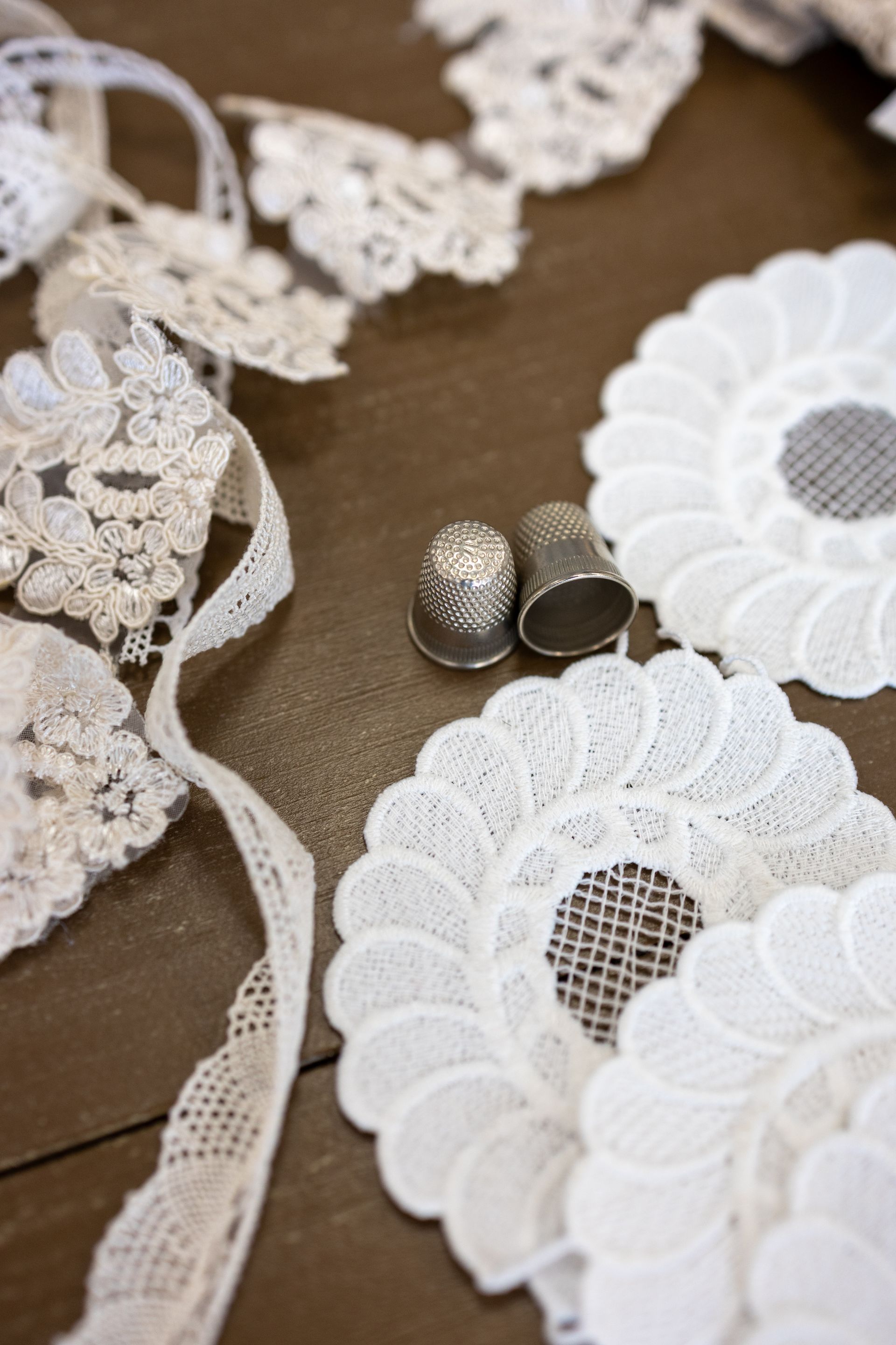 Nastri di pizzo bianco, decorazioni floreali e due ditali d'argento su una superficie di legno.