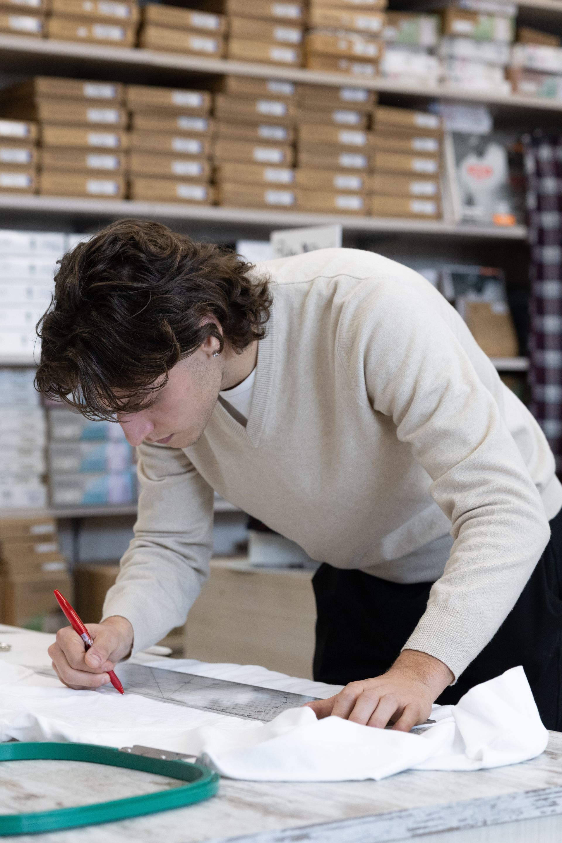 Persona con maglione leggero che disegna su un tessuto con un righello. Telaio da ricamo e scaffali sullo sfondo.