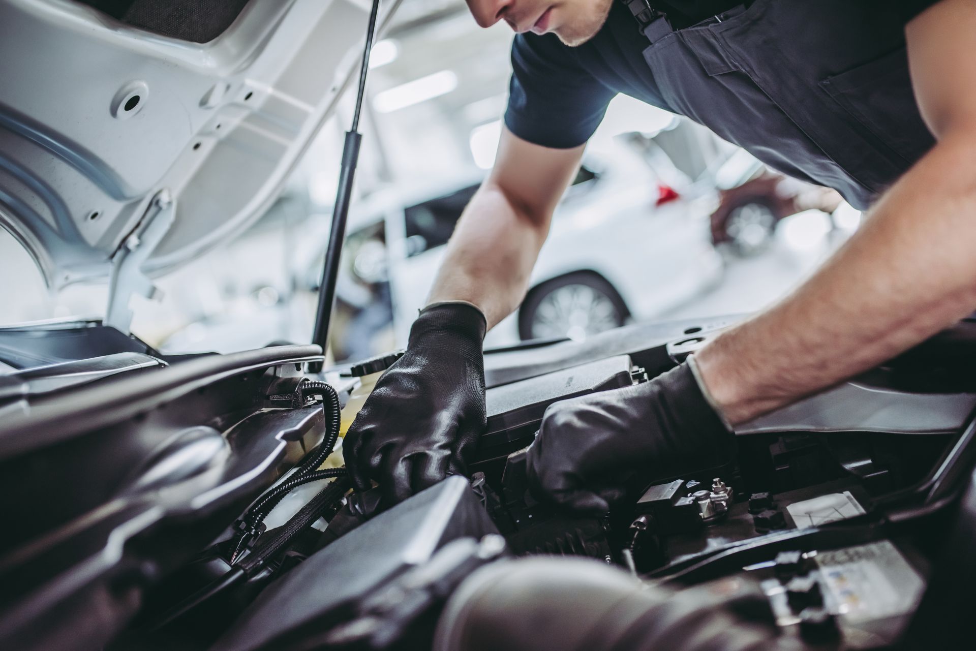 Mechanic working under a car hood in a garage, wearing black gloves and a dark shirt.