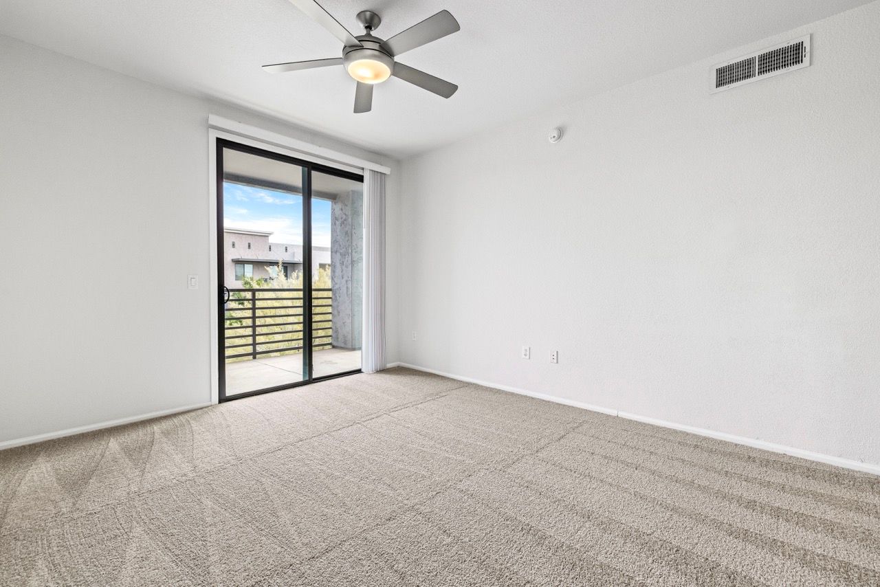 Bright living room with beige carpeting, sliding glass door to balcony, and a ceiling fan.