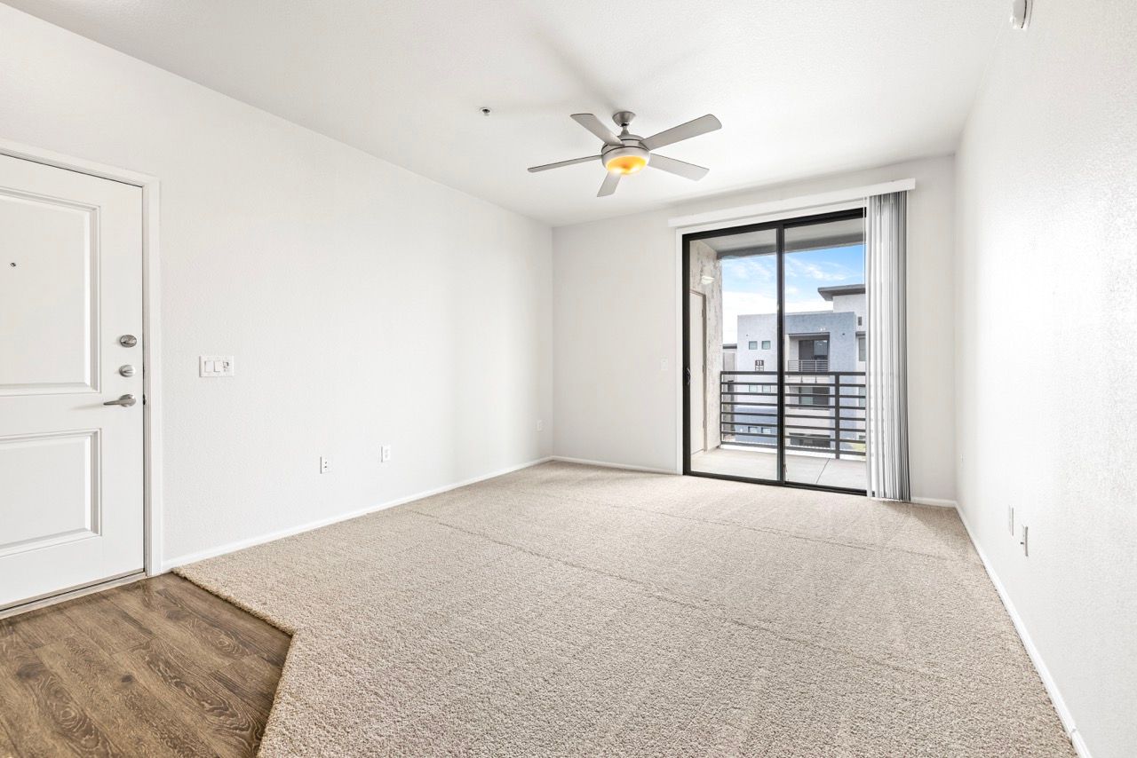 Bright living room with sliding glass door to balcony, ceiling fan, and beige carpet.