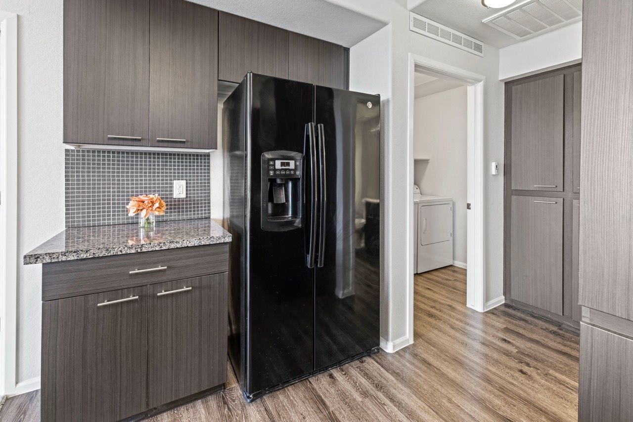 Kitchen with dark wood cabinets, granite countertop, black refrigerator, and laundry area visible through doorway.