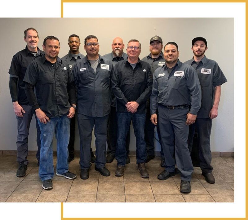 Group of mechanics, mostly men, in dark shirts and gray pants, smiling, standing in a workshop. | Arizona Engine Rebuilding