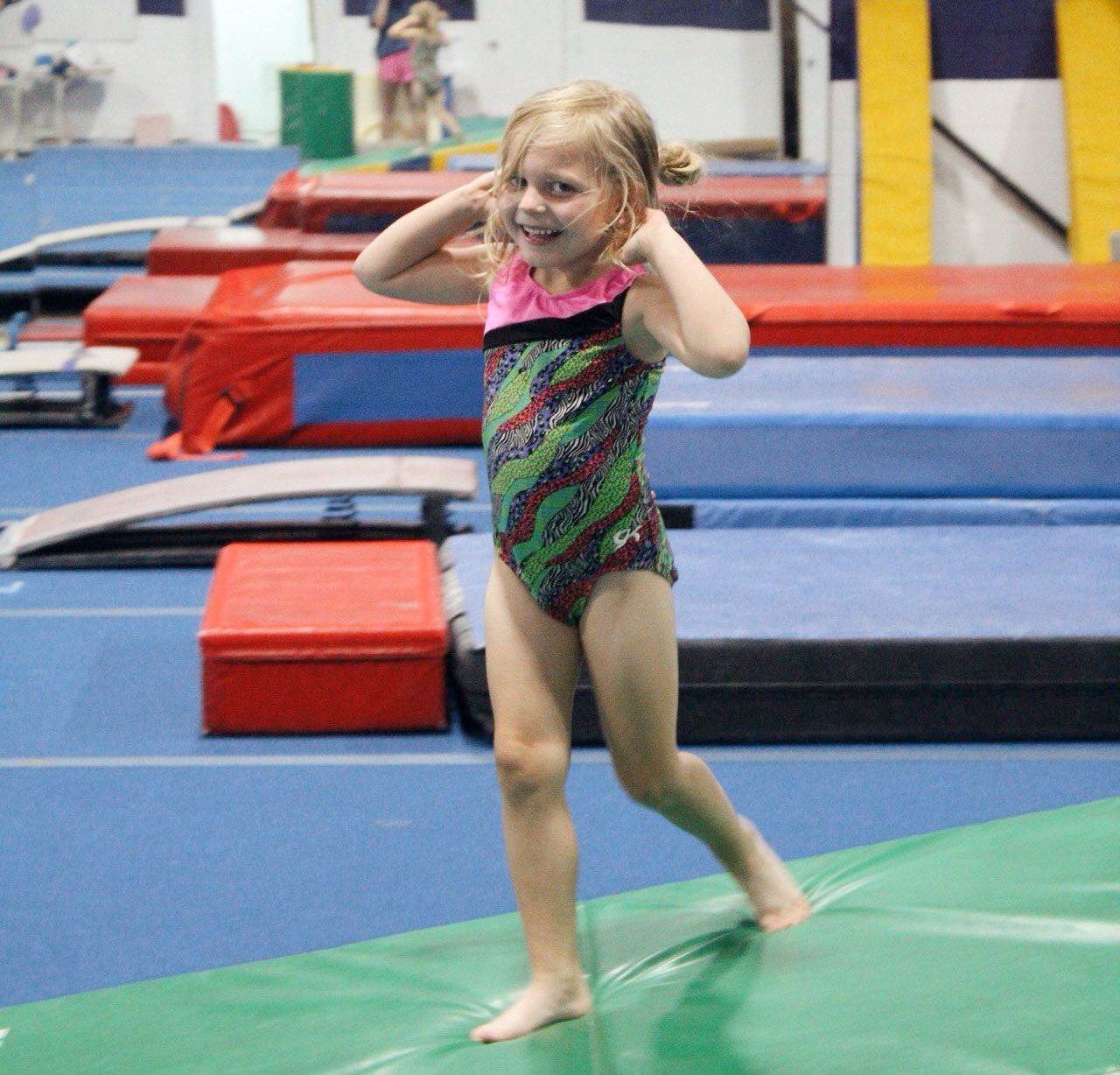 A child in a patterned leotard smiling while balancing on a green gymnastics mat in an indoor gym.