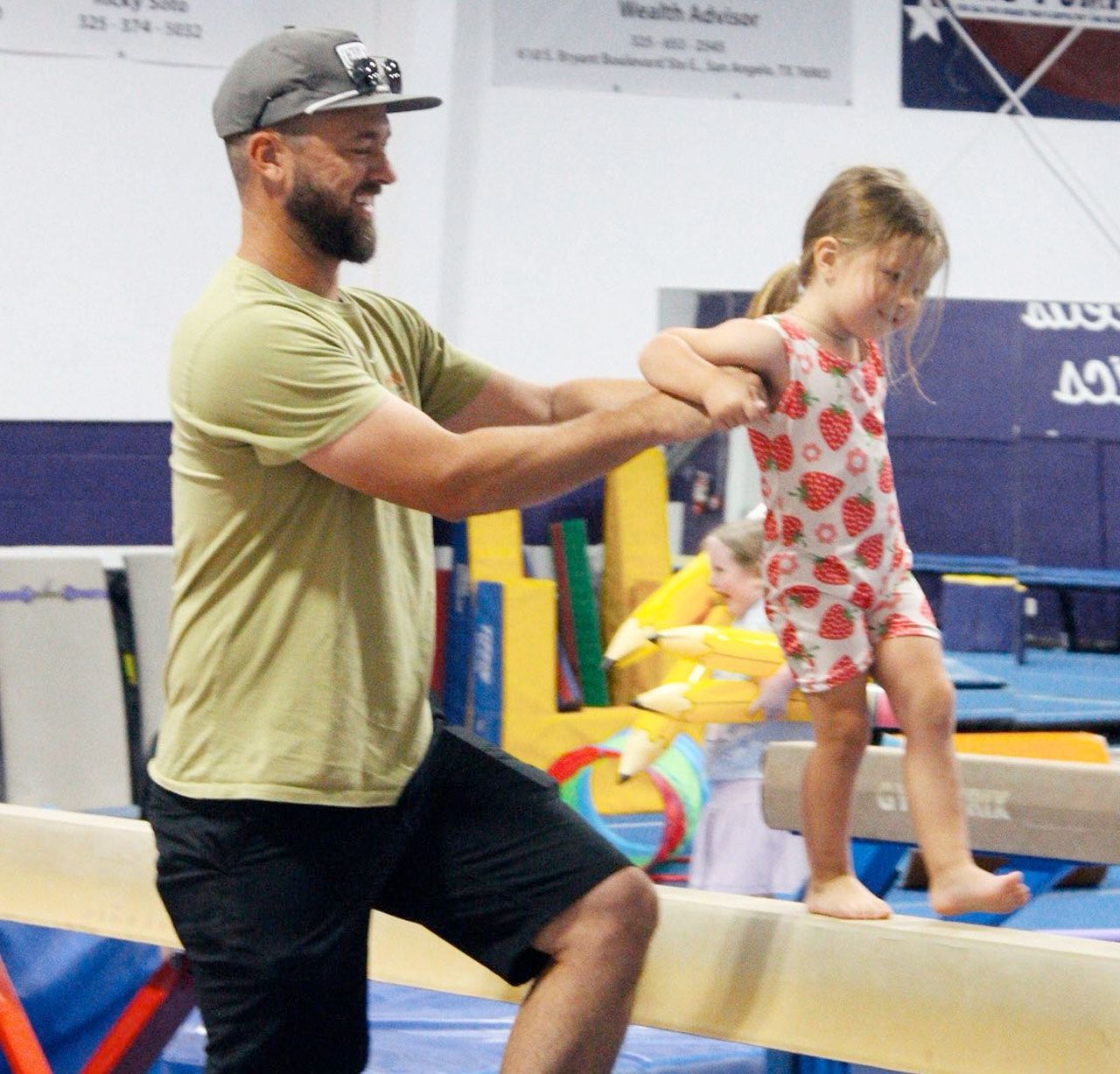 A coach in a green t-shirt and baseball cap assists a young child in a strawberry-print leotard on a gymnastics beam.