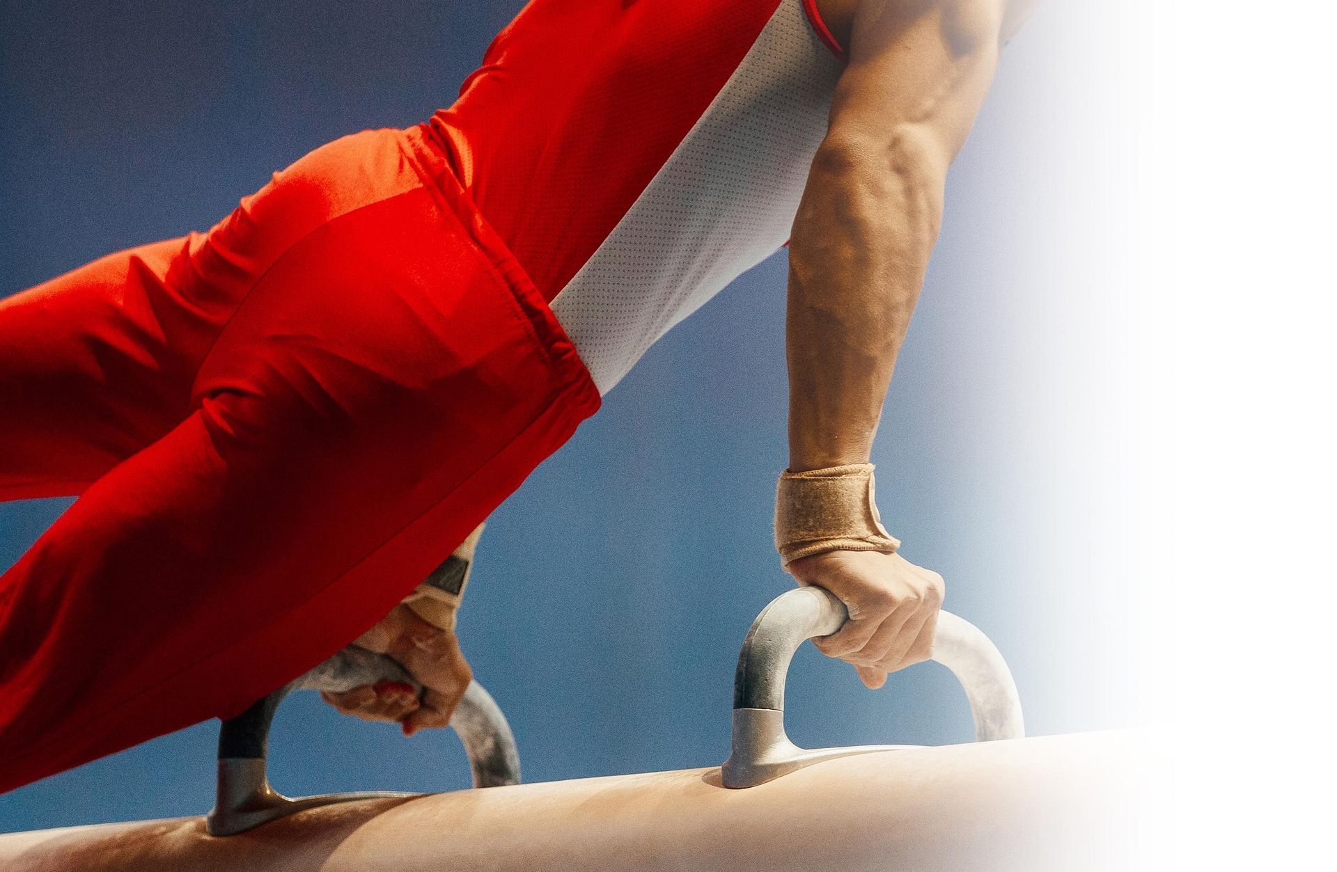 A gymnast in a red and white uniform performs on a pommel horse, hands gripped on the handles.